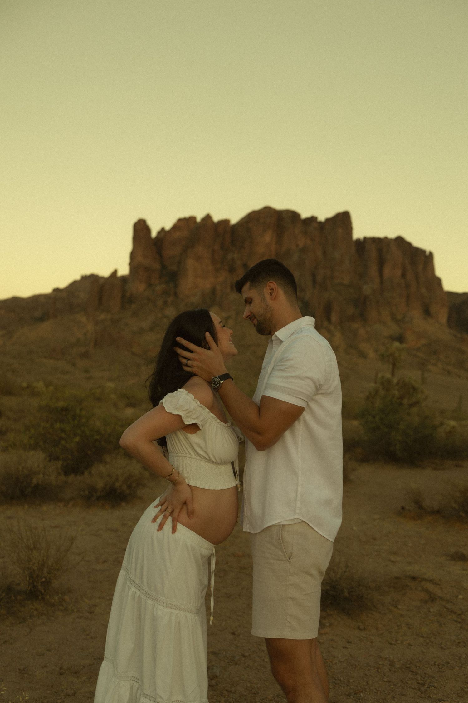 Expecting couple sharing tender moment in Sonoran Desert with rocky mountain formation at sunset.