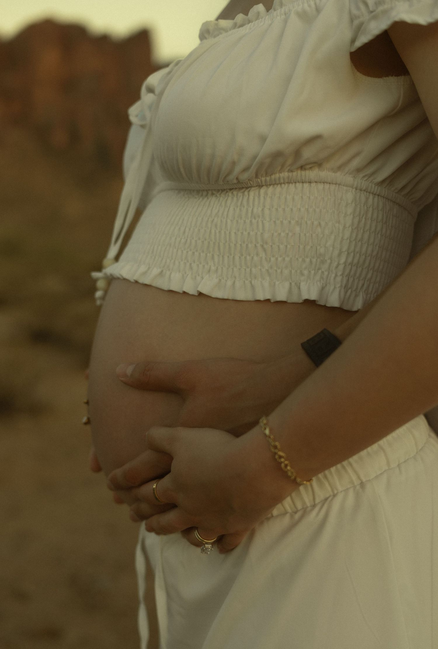 Pregnant woman in white crop top and skirt cradling baby bump outdoors, wearing gold bracelet and rings.