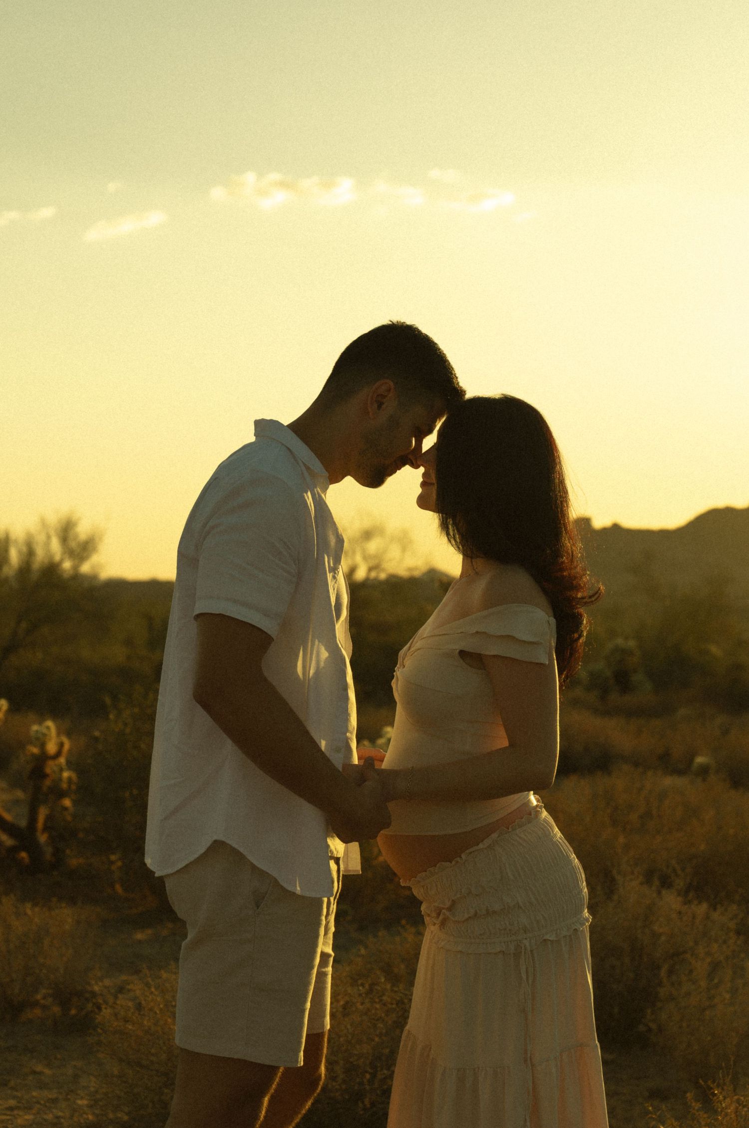 A couple shares a tender moment at sunset in a desert landscape, touching foreheads while holding hands over a baby bump.