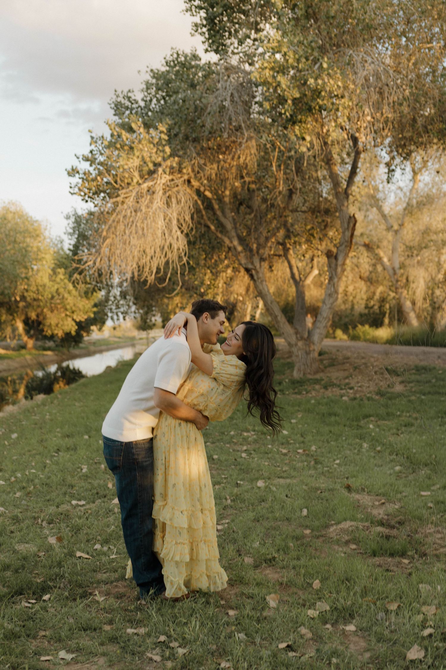 A couple embraces on a gentle slope with autumn trees in the background during sunset.