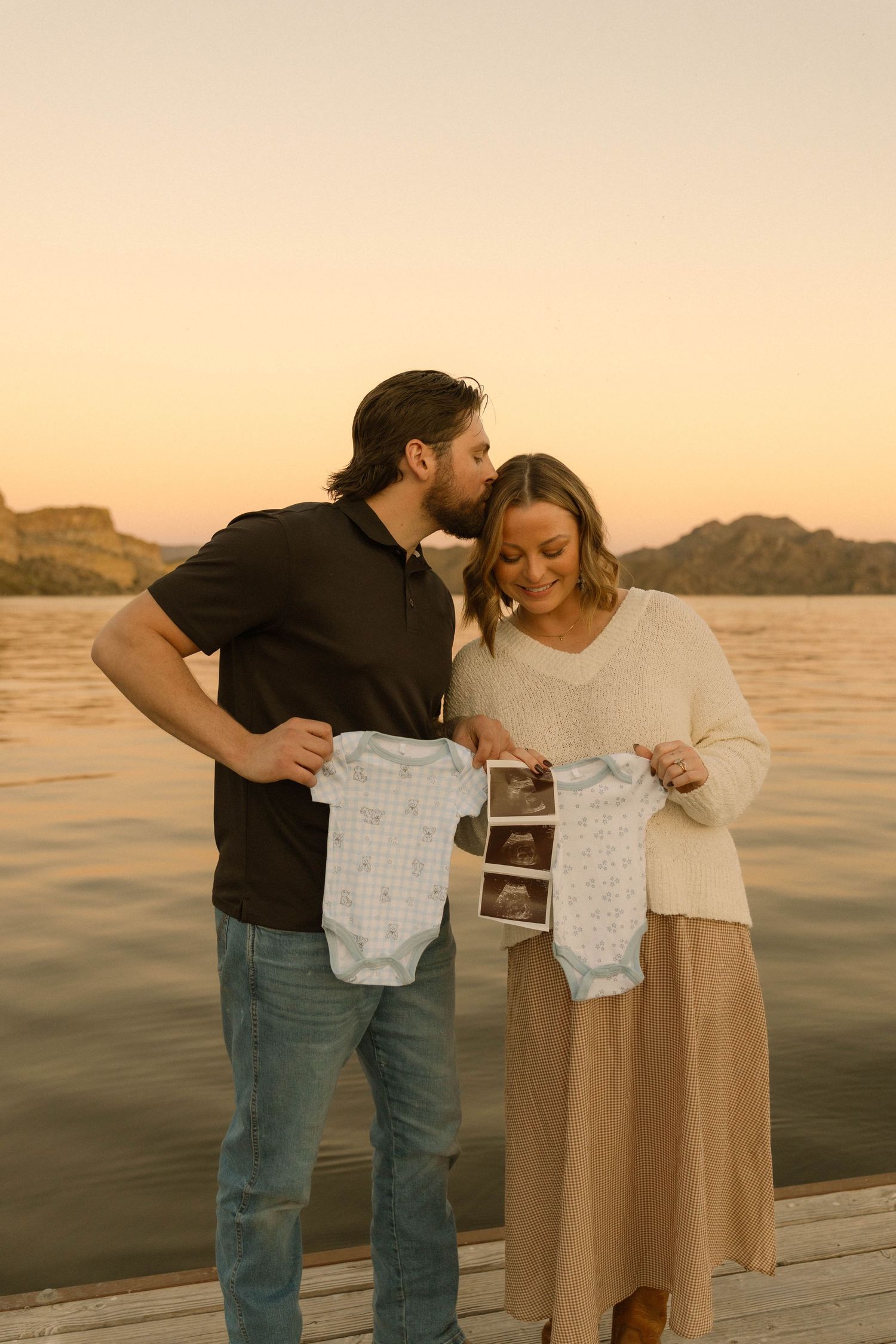 Happy couple displays baby clothes and sonogram photos on lakeside dock at sunset.