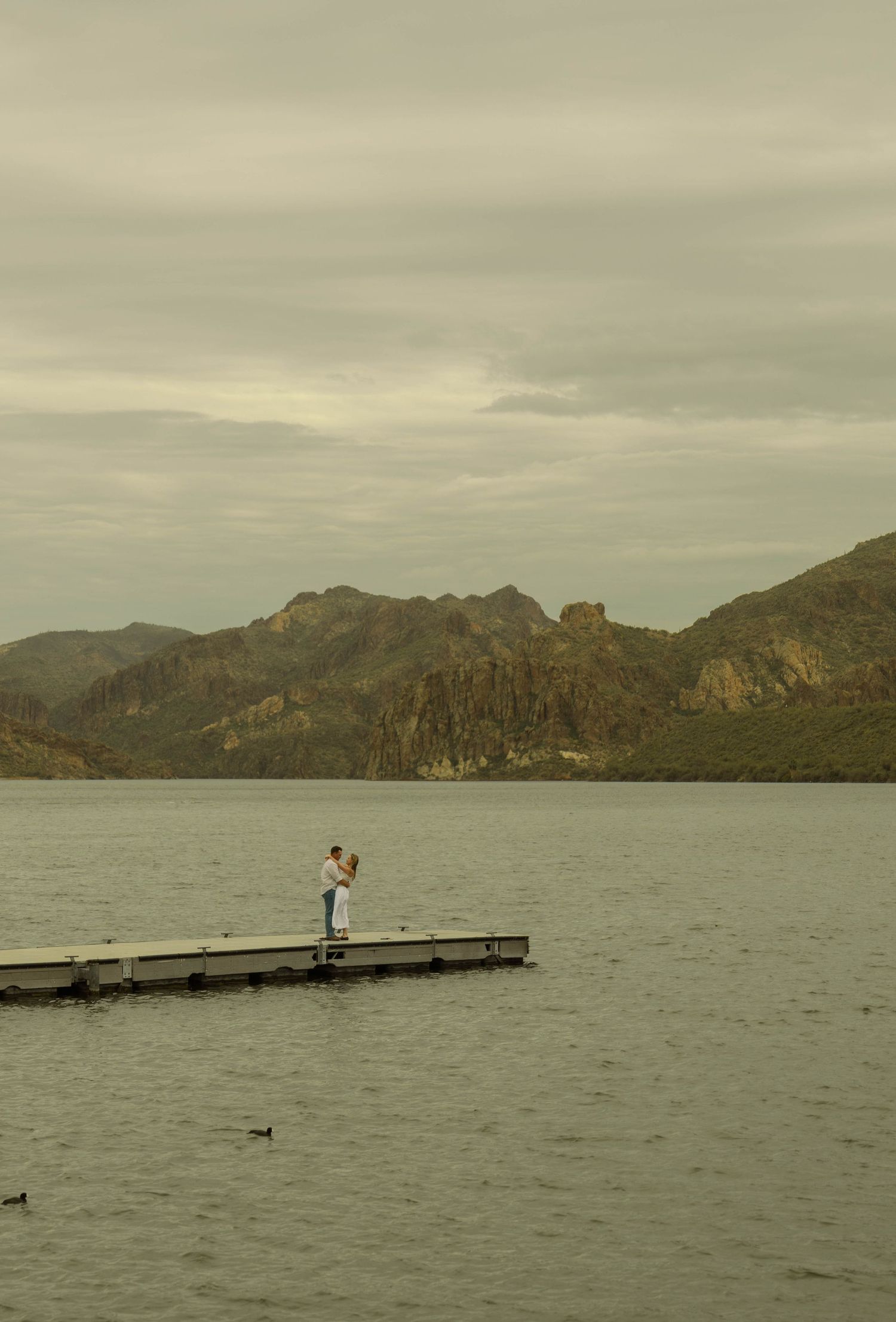 Panoramic view of a lake surrounded by rugged mountains with a wooden dock and distant figures in the water.