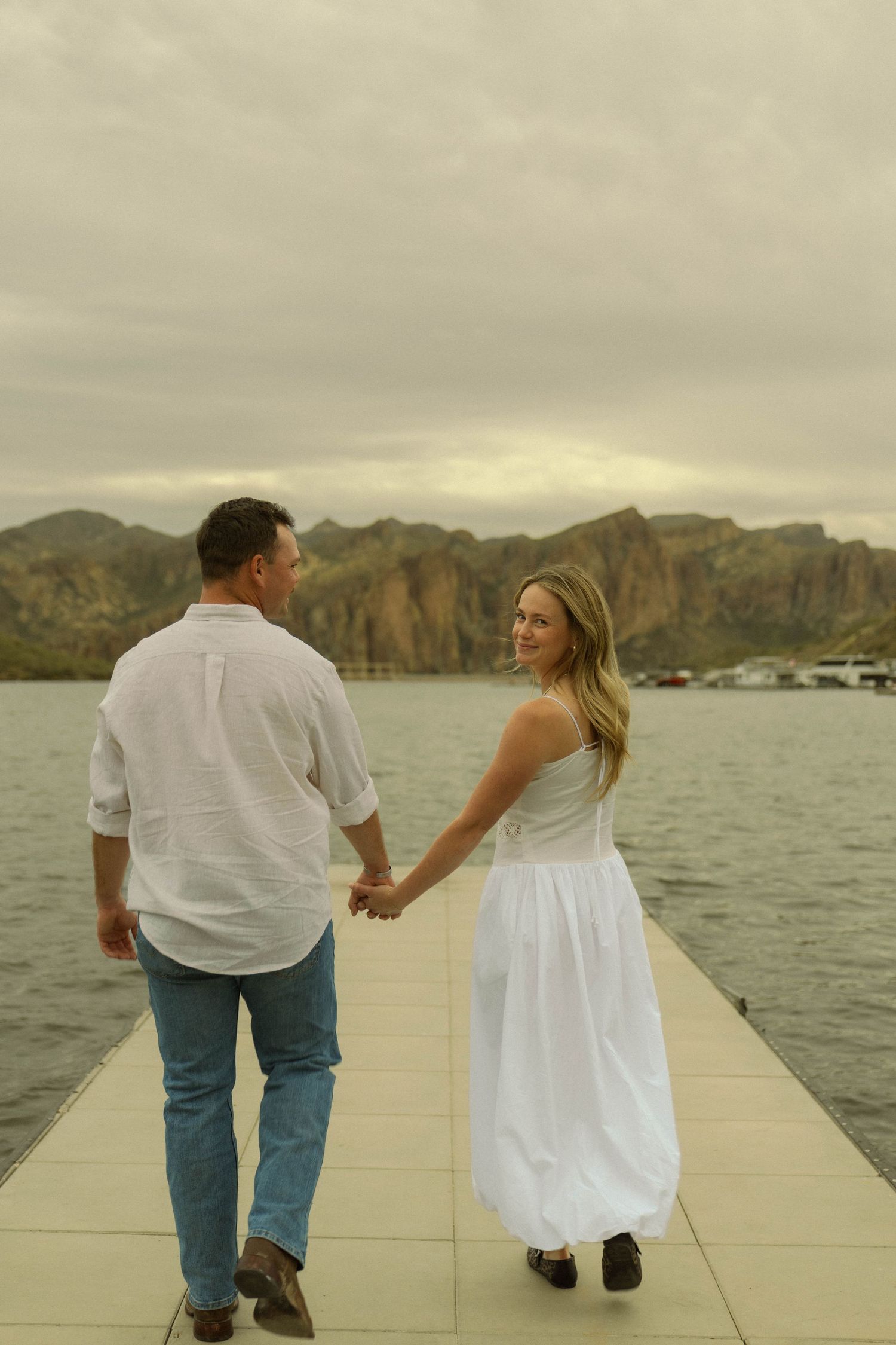 A couple sits on a dock overlooking a scenic lake surrounded by mountains at sunset.