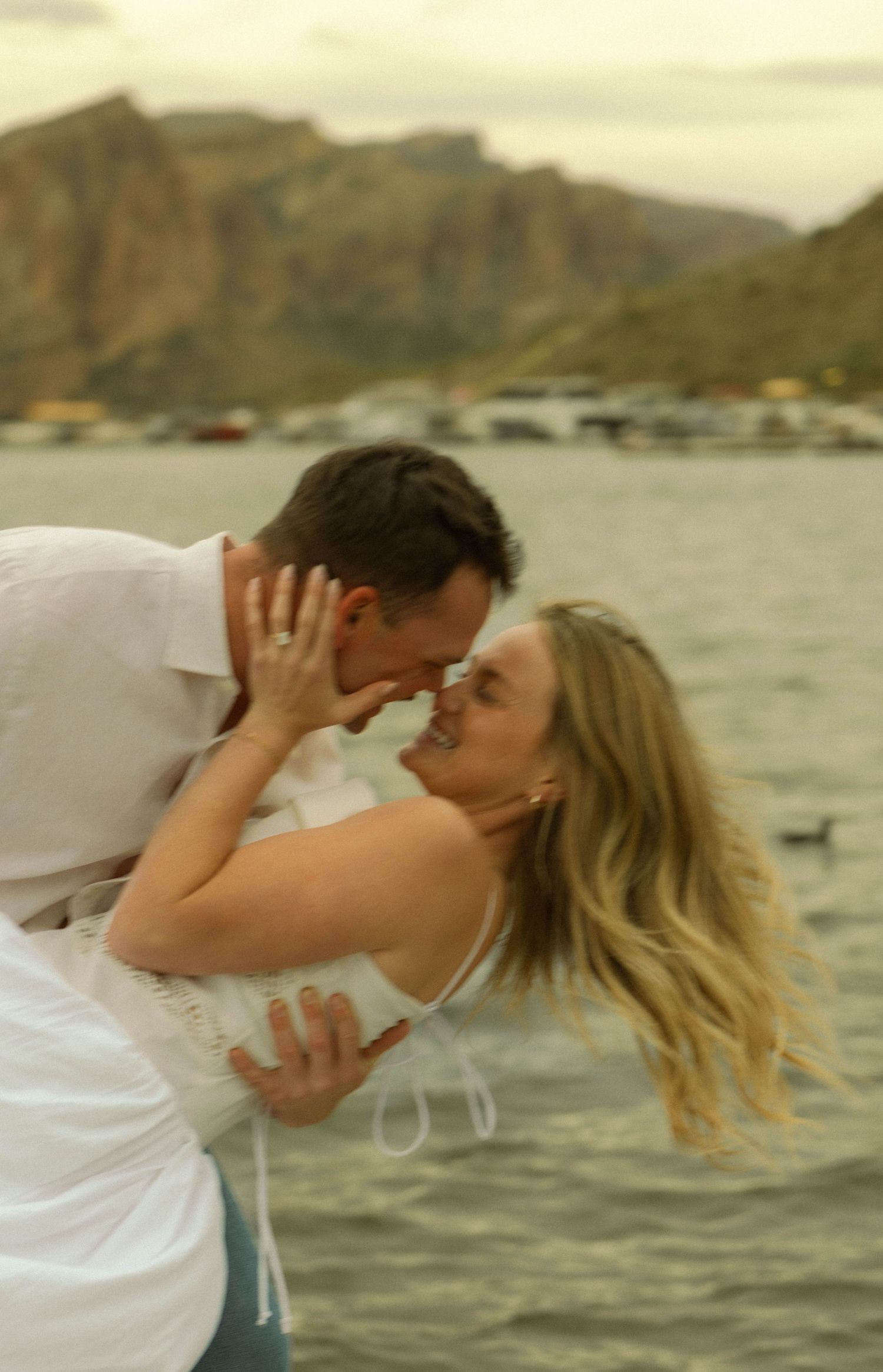 A playful couple dips and embraces near the ocean with dramatic cliffs in the background.