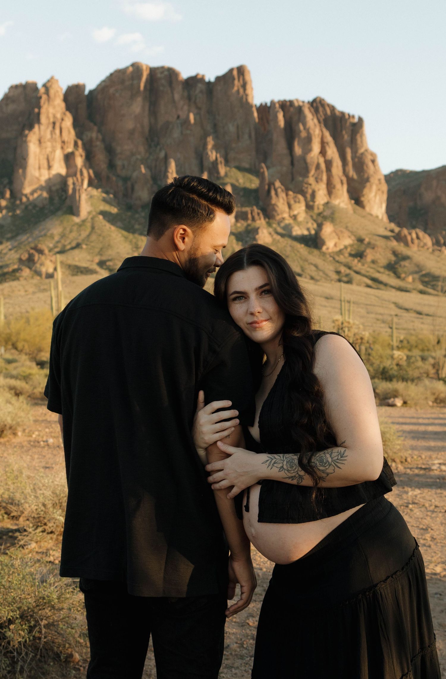 A couple embraces tenderly against a backdrop of rugged desert mountains during a golden hour photoshoot.