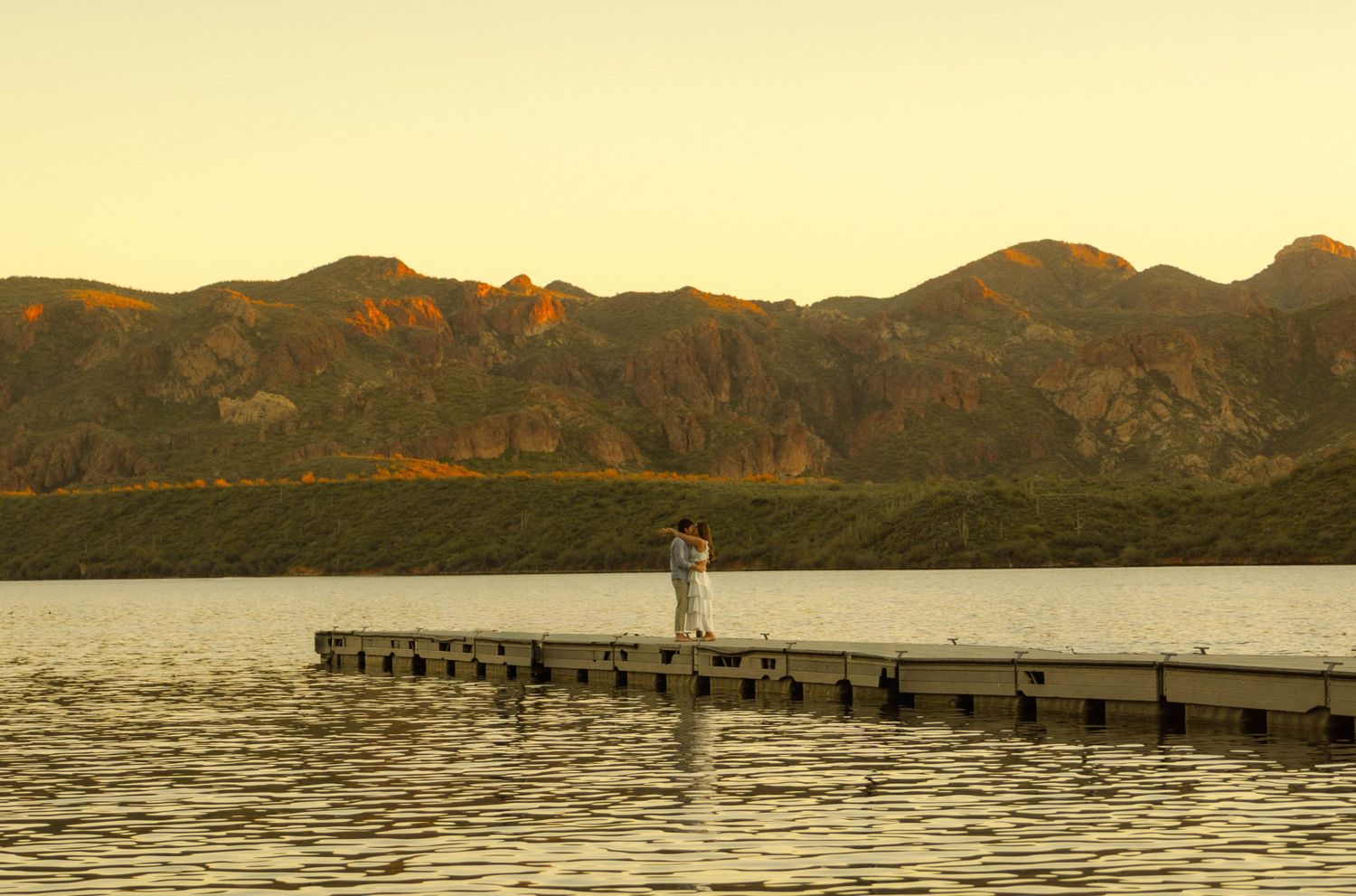 Scenic mountain lake landscape at sunset with wooden pier extending over calm waters and rugged peaks in background.