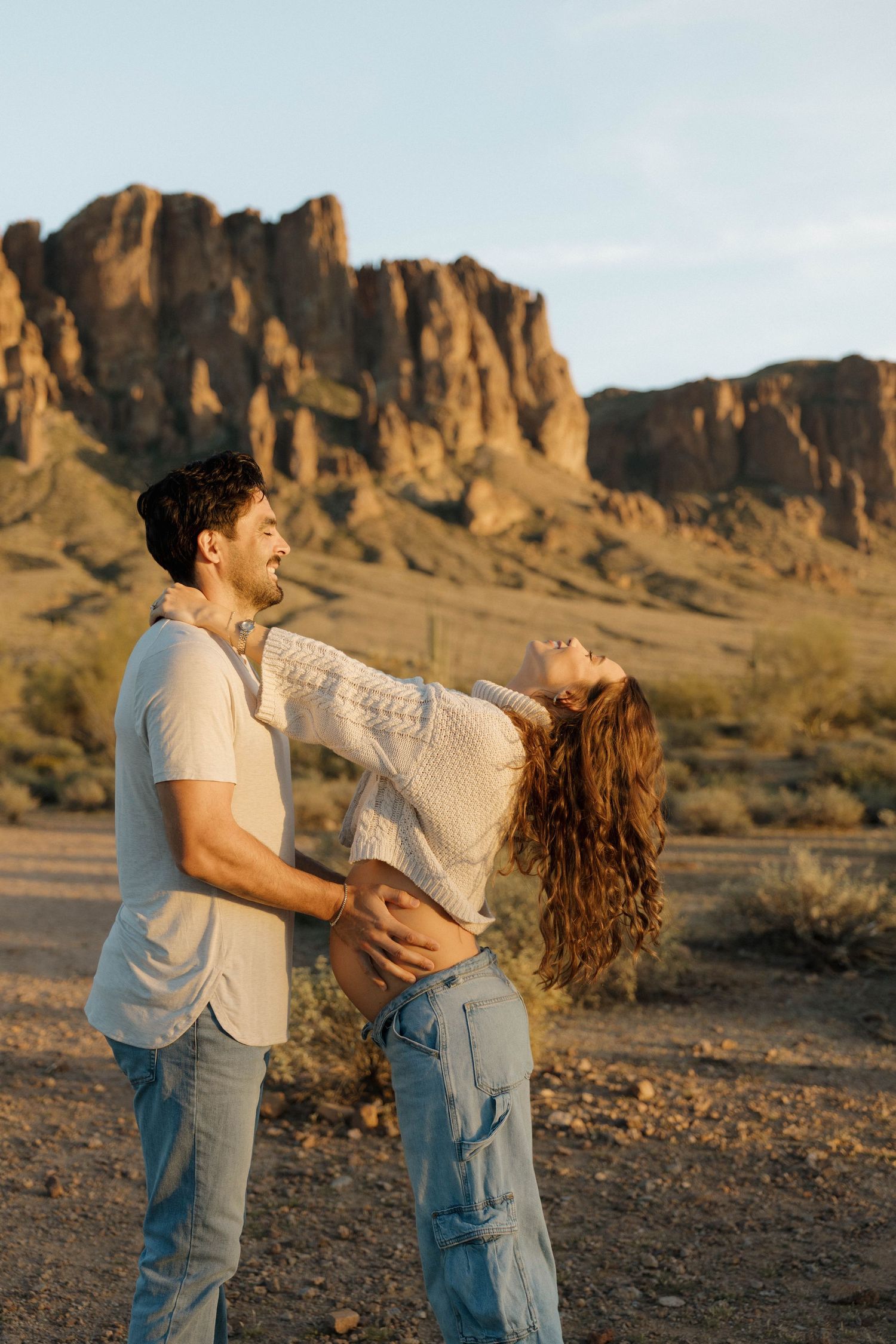 A couple embraces and dances playfully in front of scenic desert rock formations at golden hour.