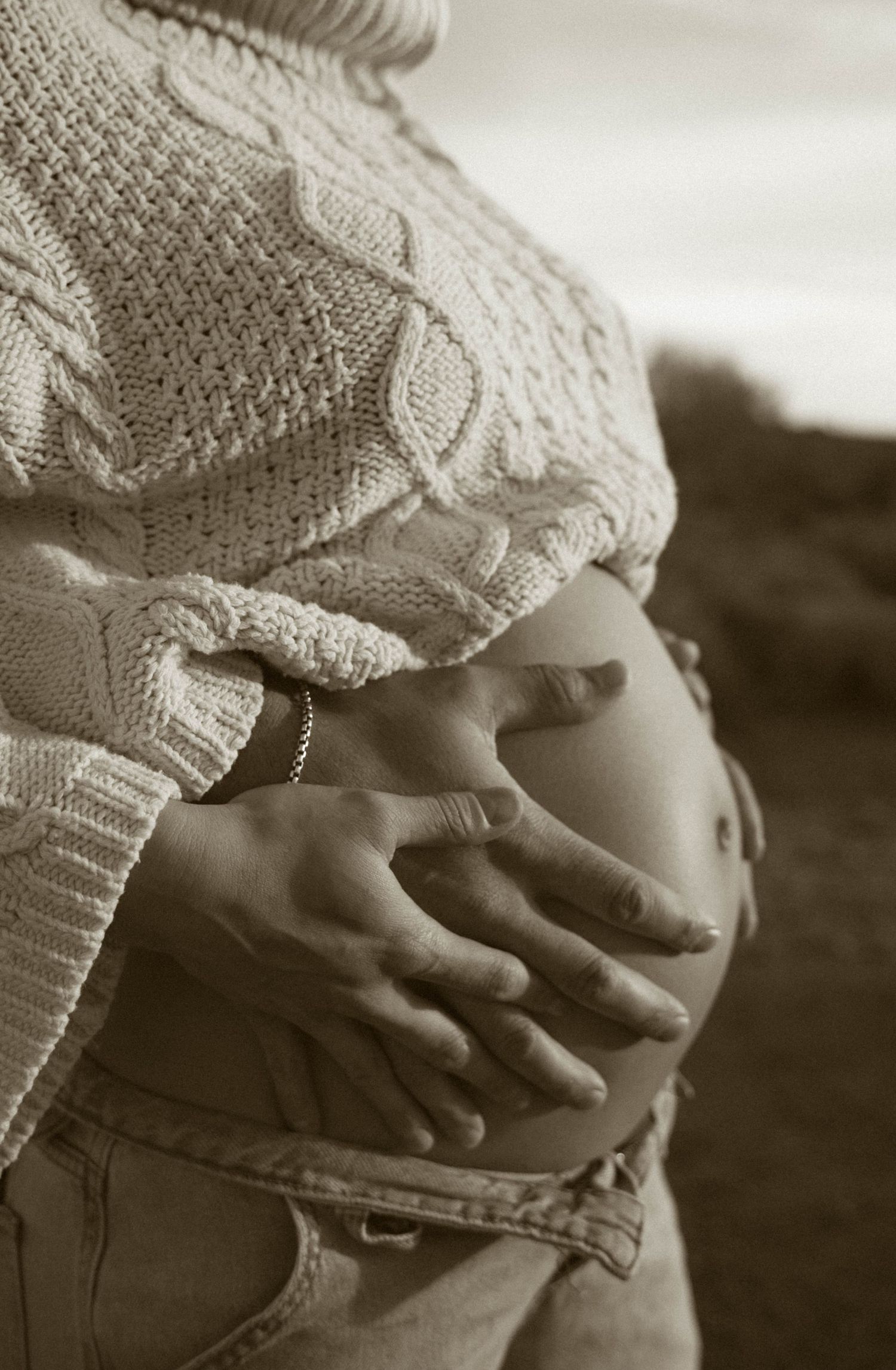 A black and white photograph shows hands gently cradling a pregnant belly in a knit sweater.
