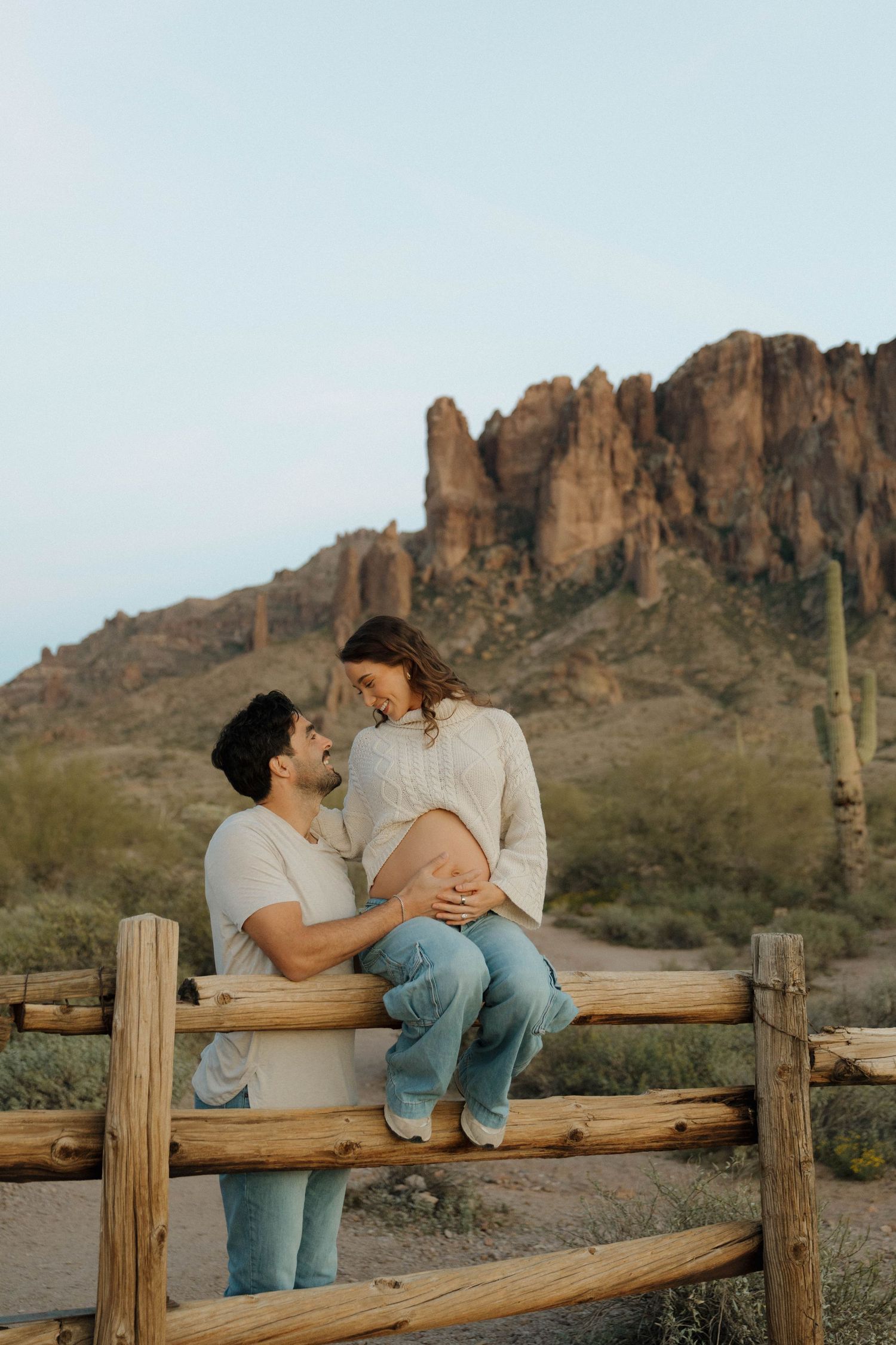 A couple shares an intimate moment on a wooden fence with the Superstition Mountains and desert landscape in the background.
