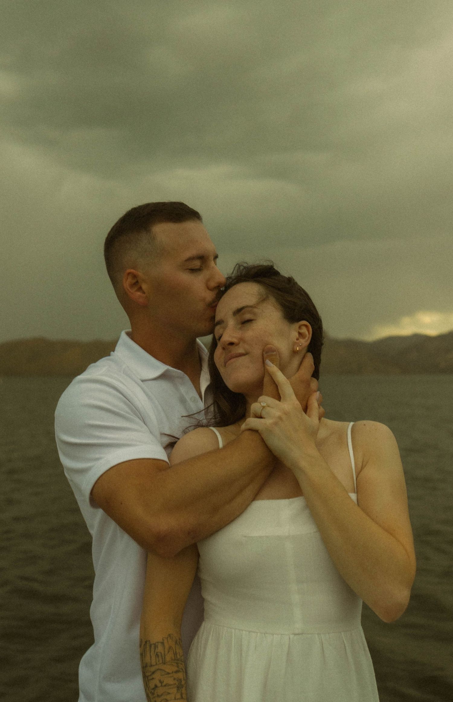 A couple in white outfits share an intimate moment against a moody, cloudy seascape backdrop.