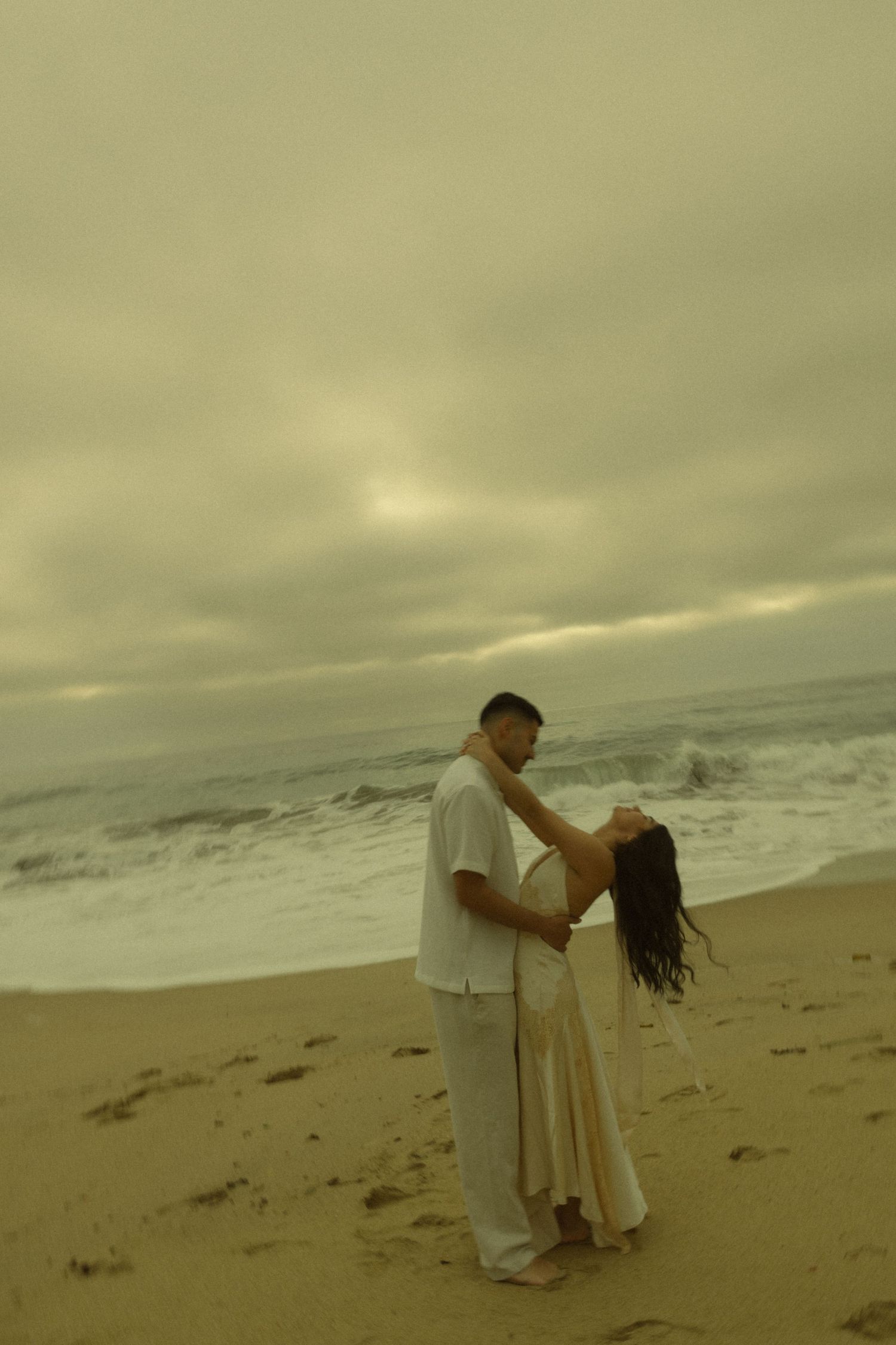 Romantic couple embracing on a sandy beach with dramatic overcast skies and ocean waves in the background.
