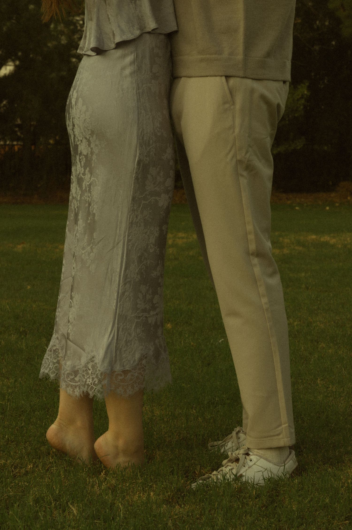 Couple standing close together on grass, woman in lace dress barefoot, man in cream trousers and white sneakers.