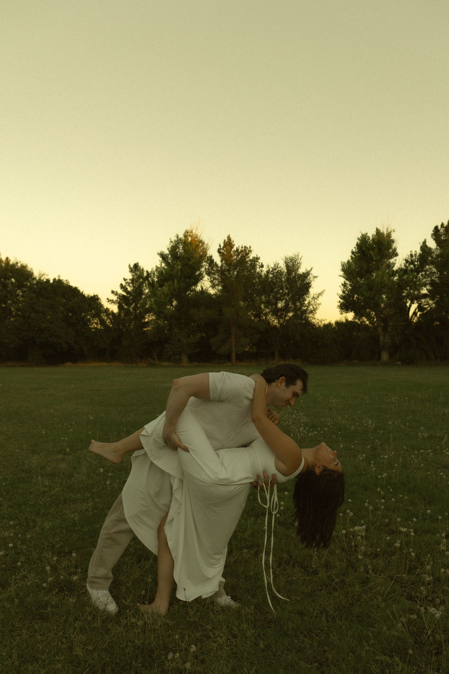 Couple sharing a romantic dip dance pose in an open green field during a warm golden sunset.