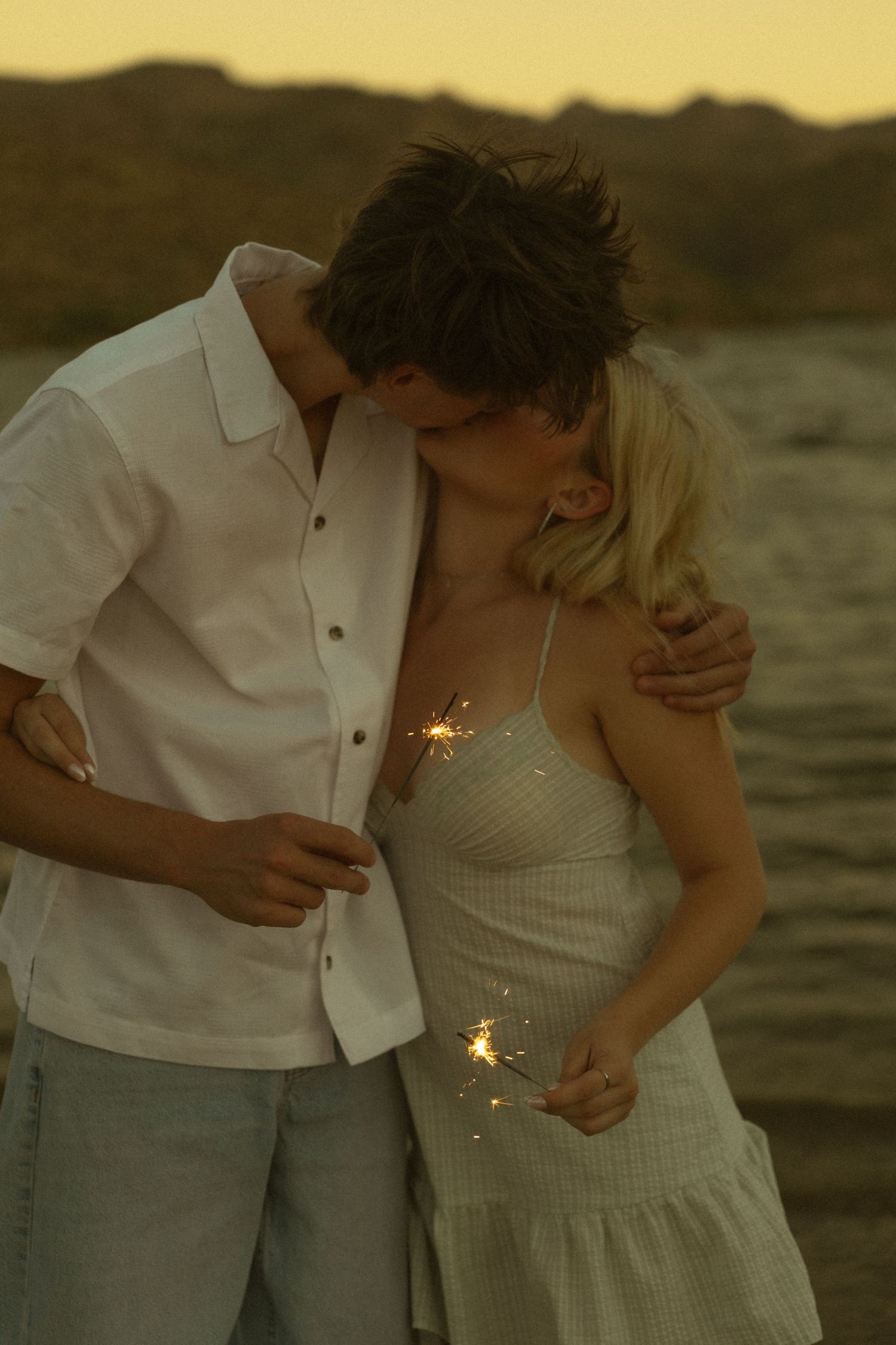 Couple kissing at sunset on beach holding sparklers, romantic golden hour photography.