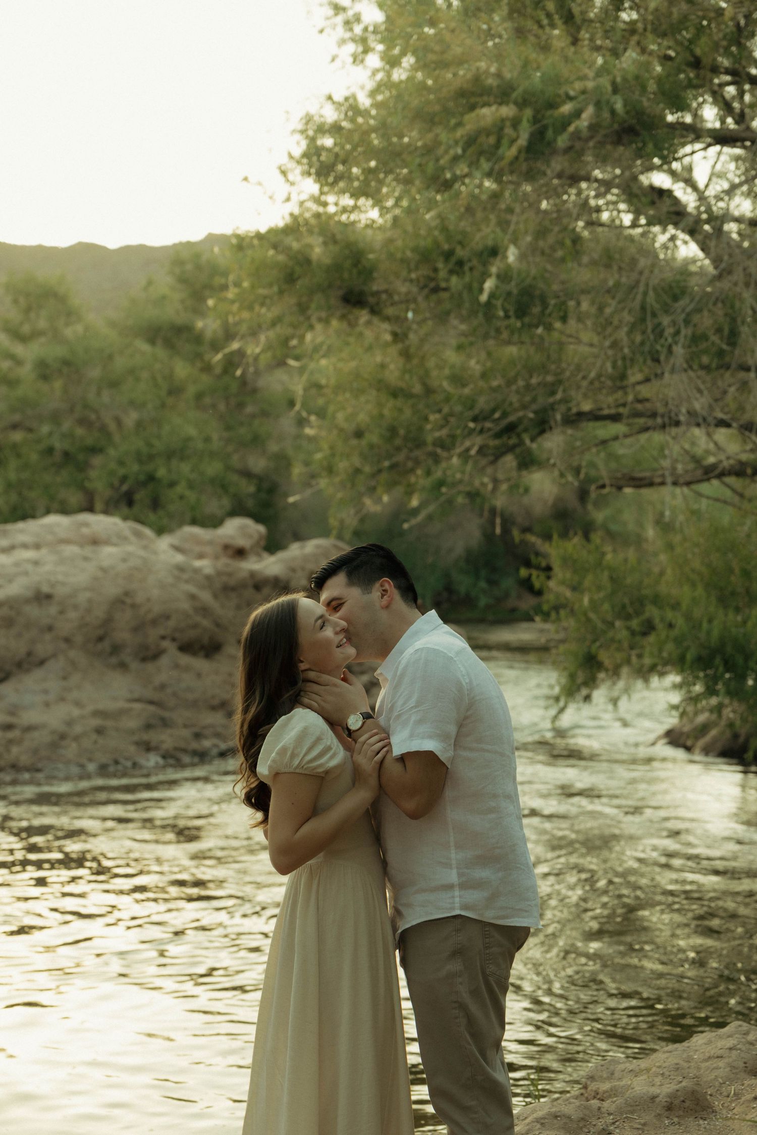 Couple sharing a romantic moment by a serene river surrounded by lush green trees during golden hour.