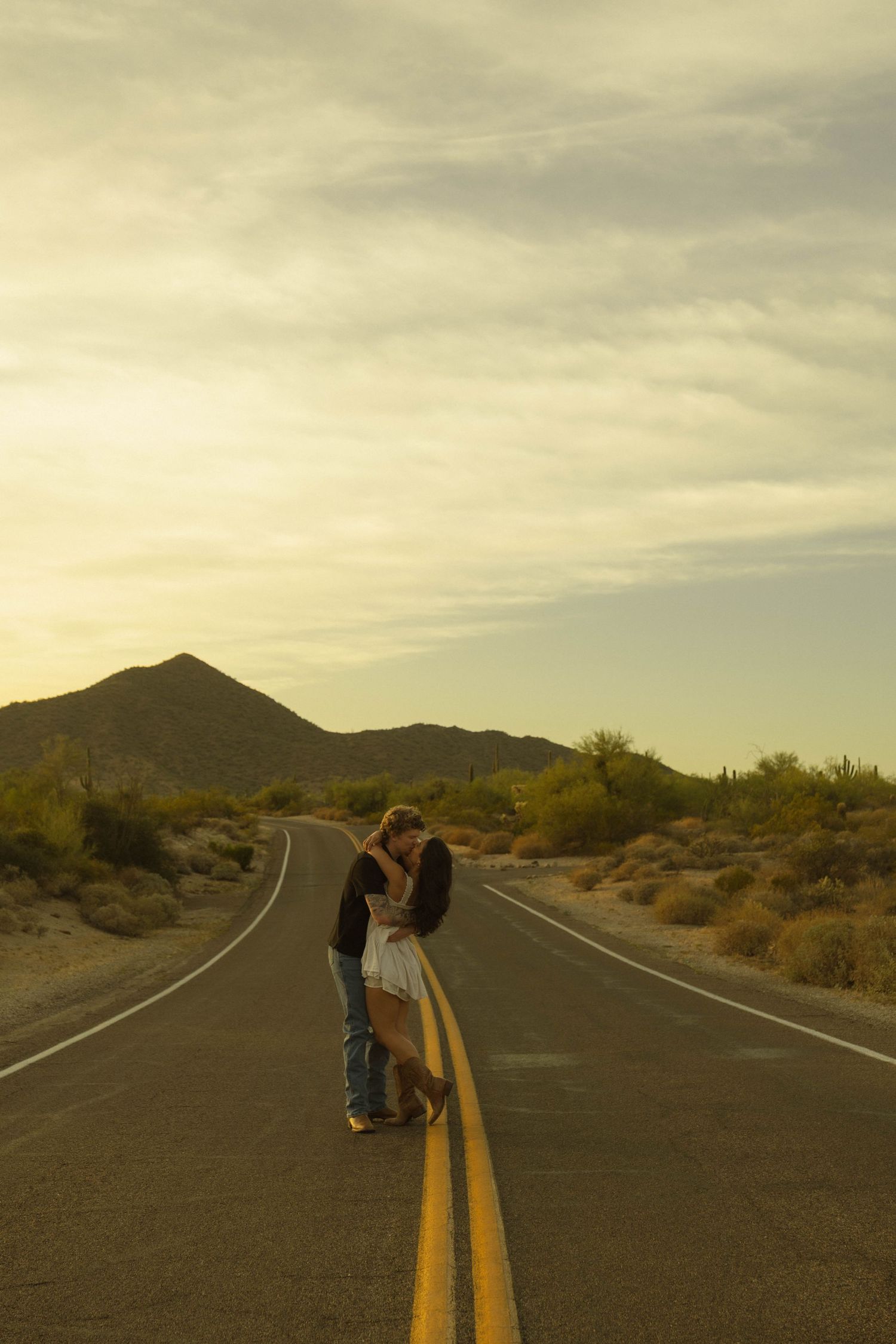 Couple kissing on desert highway centerline at sunset with mountains in background.