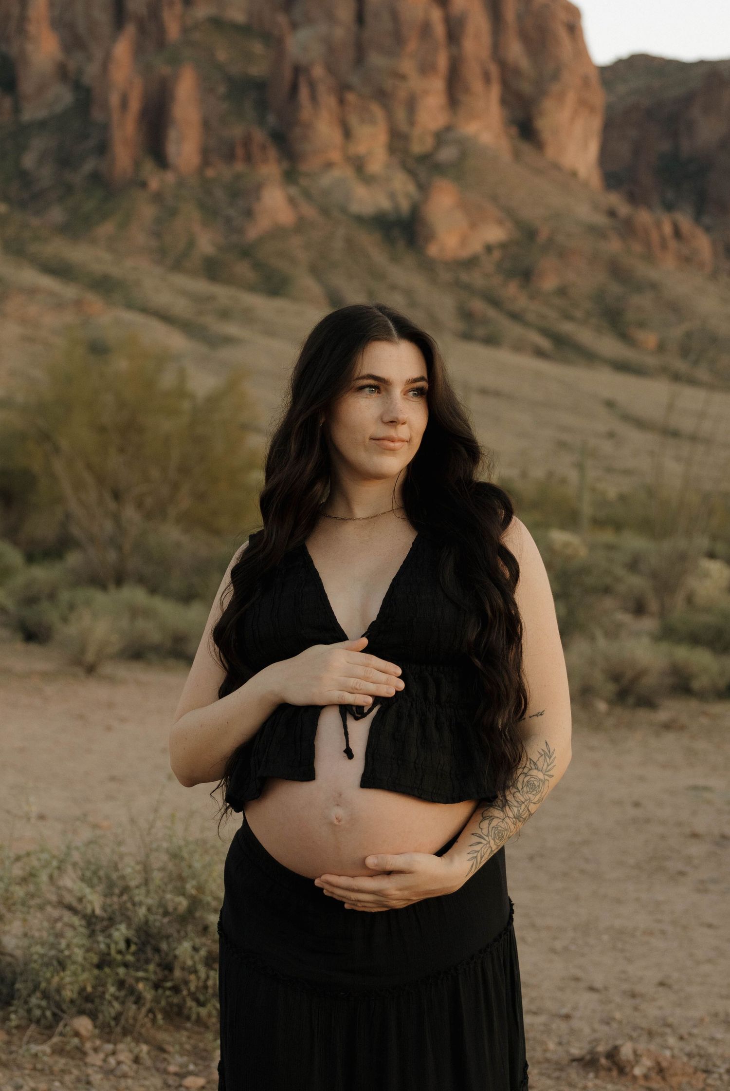 A woman in a black dress poses for maternity photos with desert mountains in the background.
