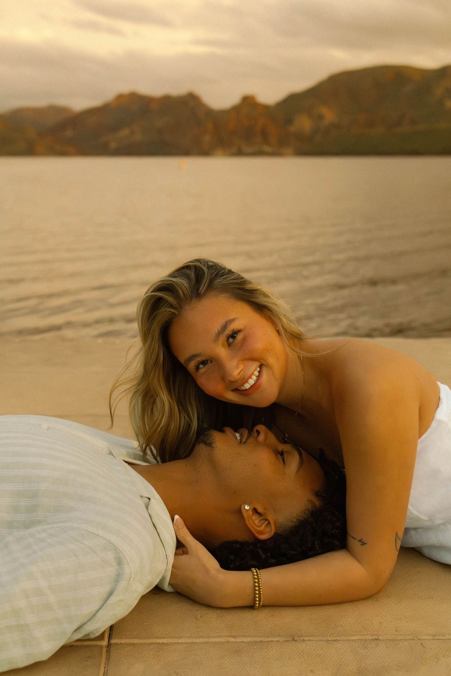 A person lying on a beach at sunset with mountains in the background, wrapped in white linens.