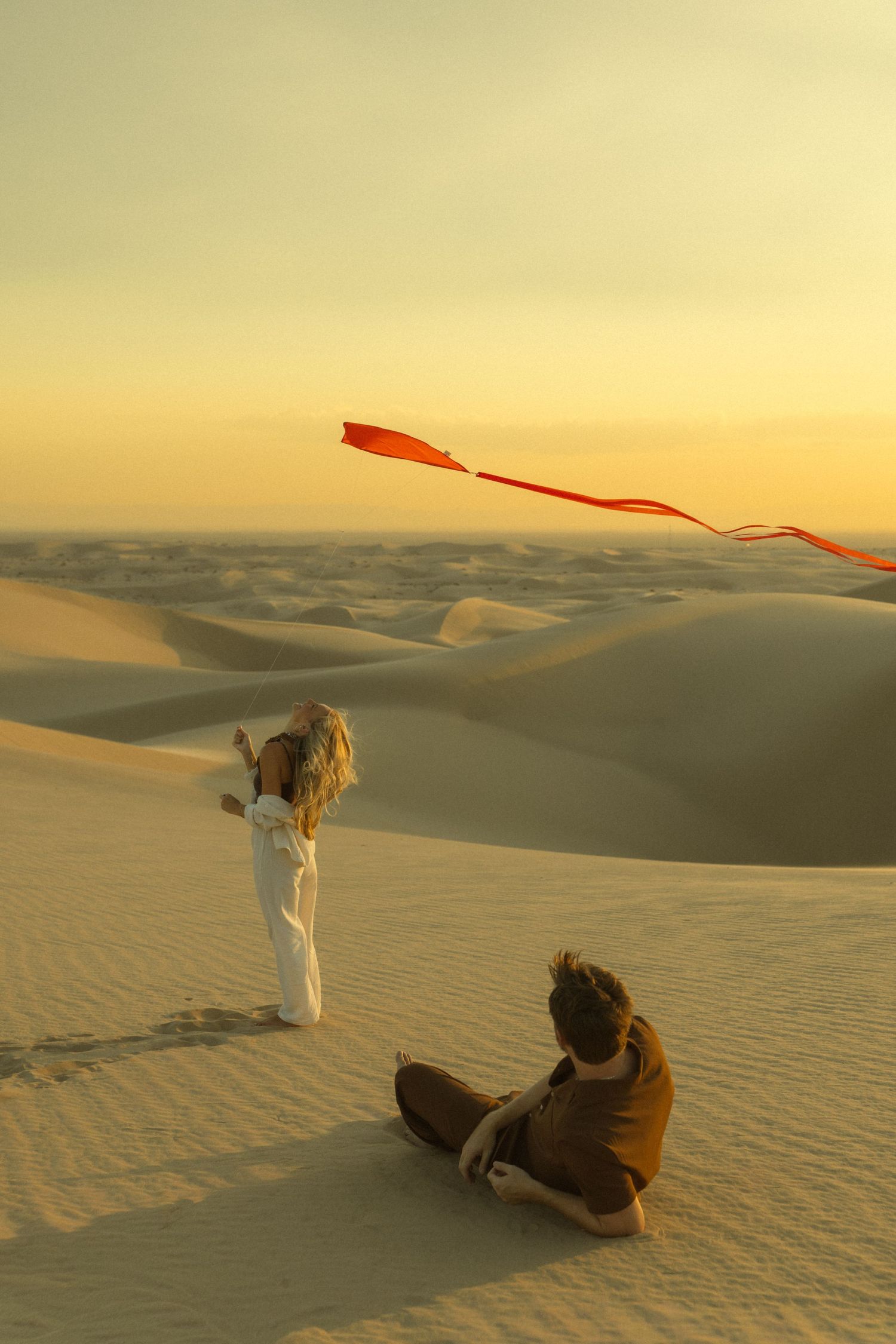 Couple enjoying a red kite flying session on expansive desert sand dunes during a warm golden sunset.