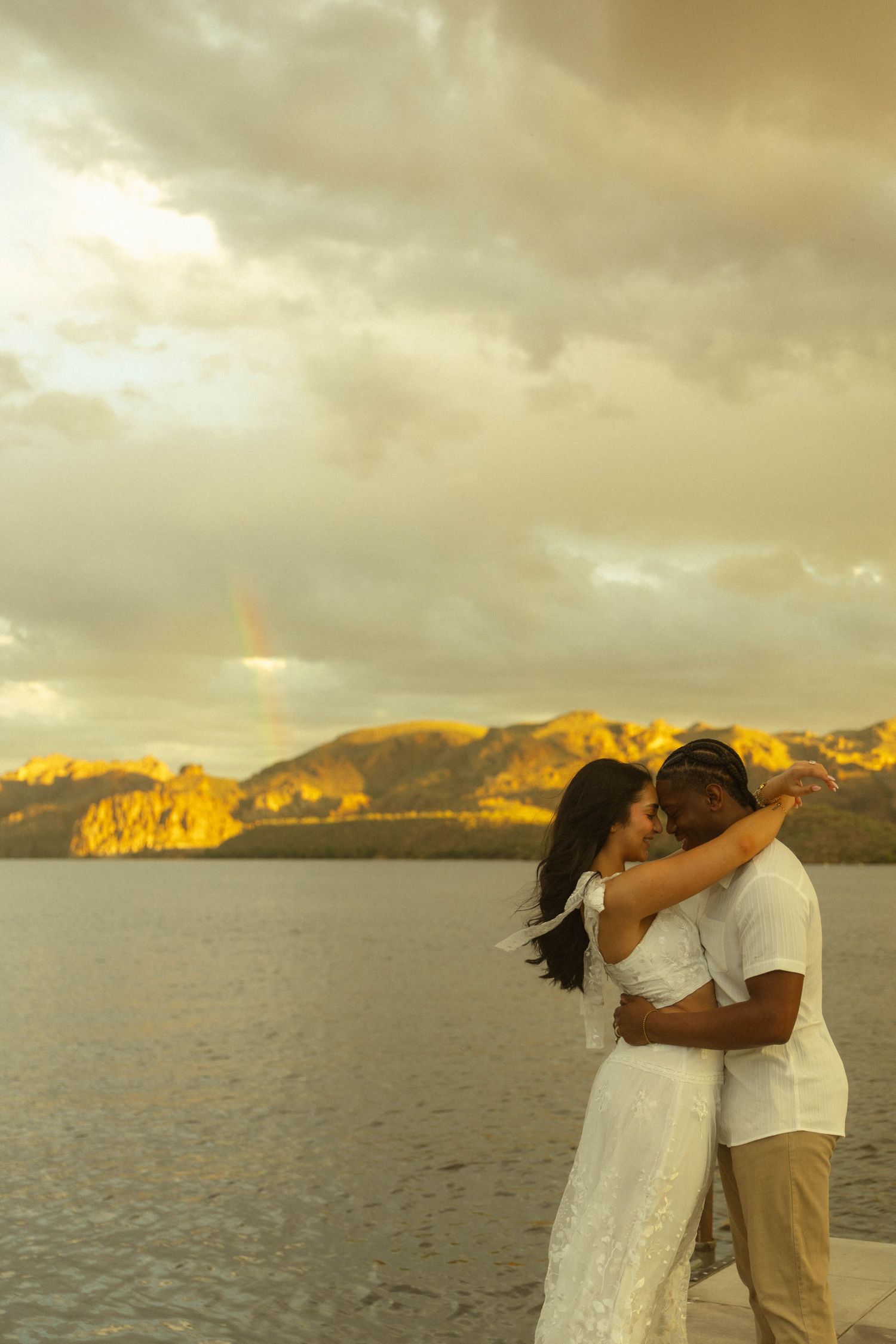 Romantic couple kissing on a lakeside dock with glowing mountains and a rainbow at golden hour.
