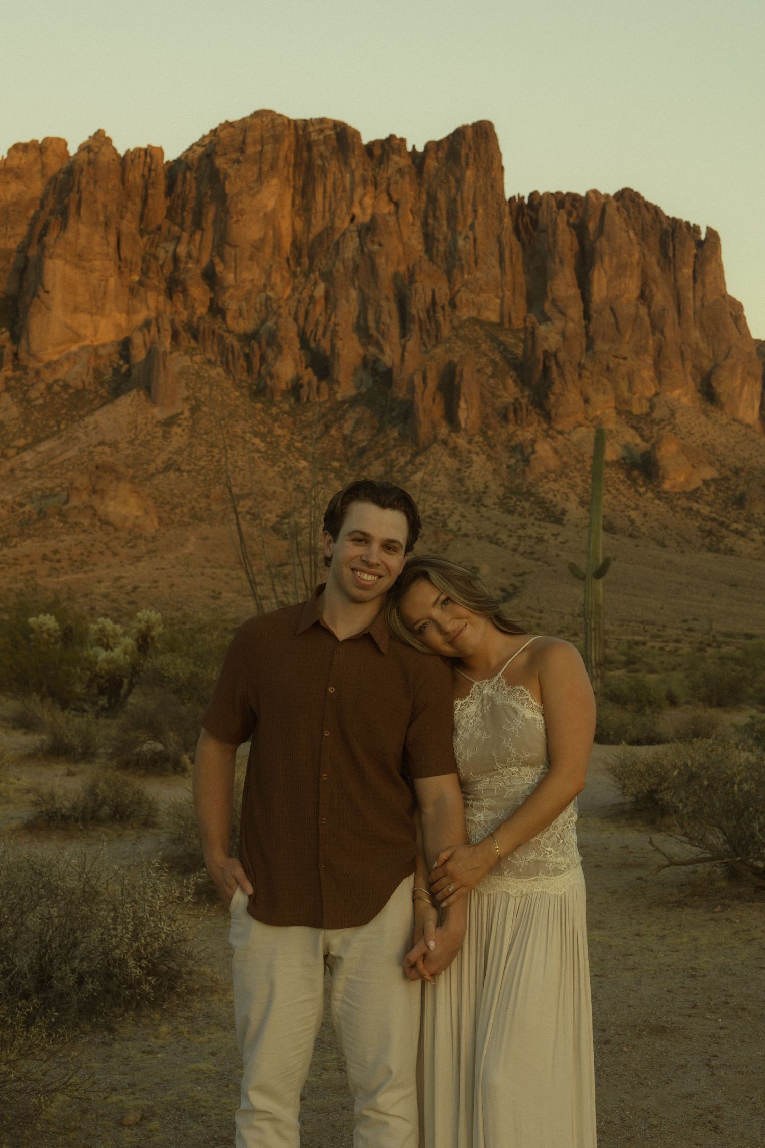 Smiling couple posing together at golden hour with the glowing Superstition Mountains in the background.