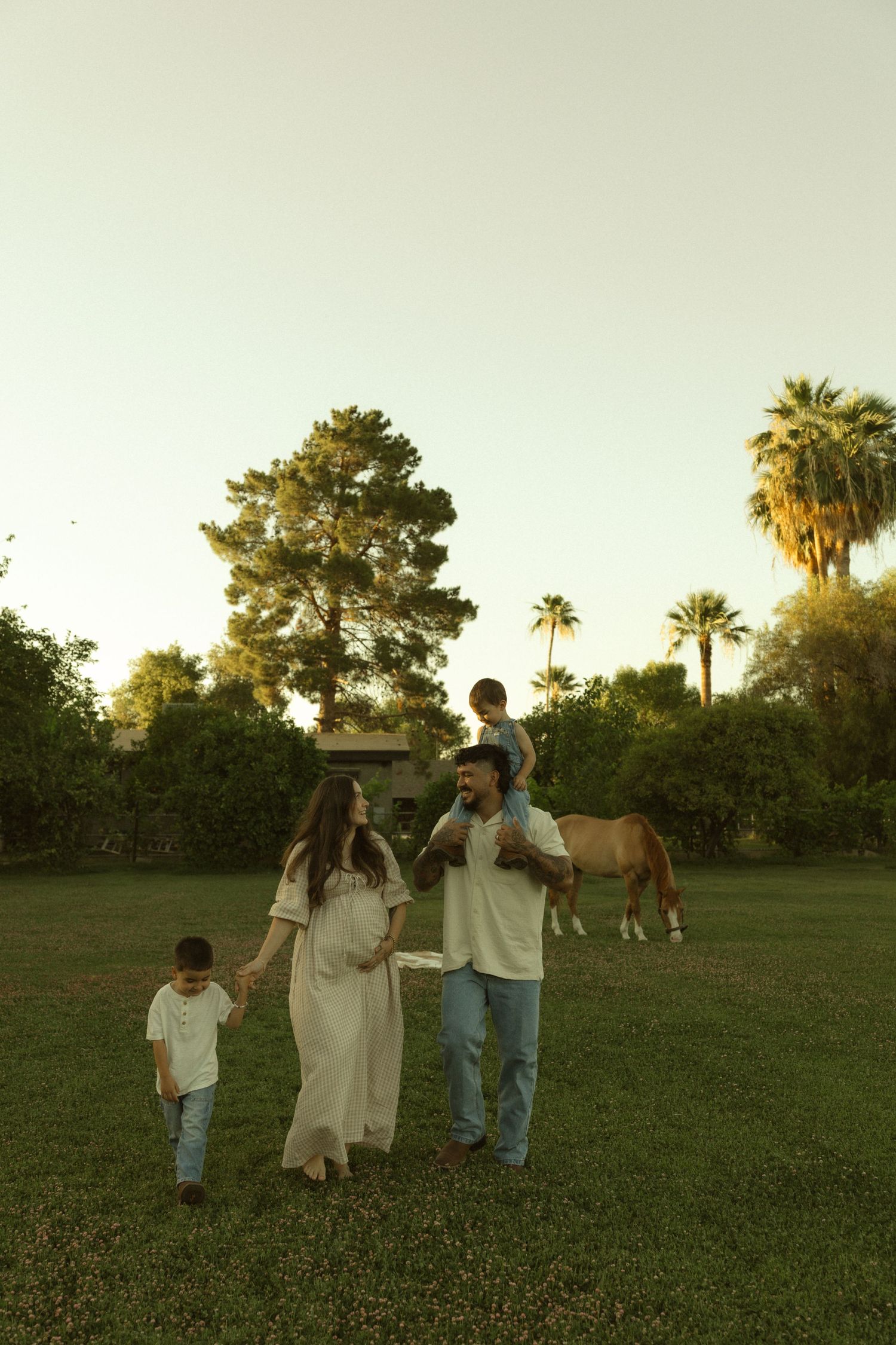 Happy family with pregnant mother, father, and two young children walking in a green field with a horse at sunset.