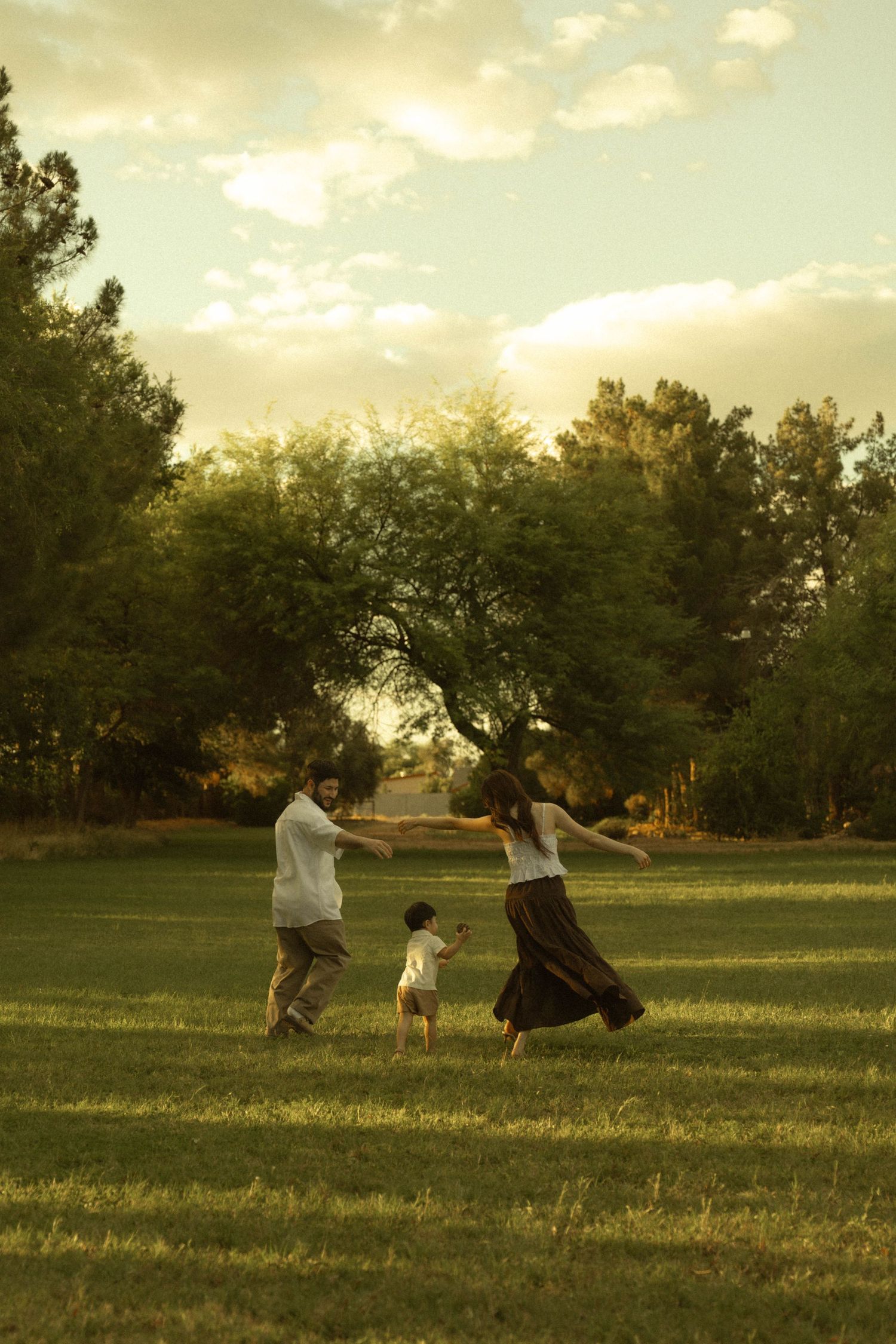 A joyful family of three plays together on a green lawn surrounded by trees during a golden sunset.