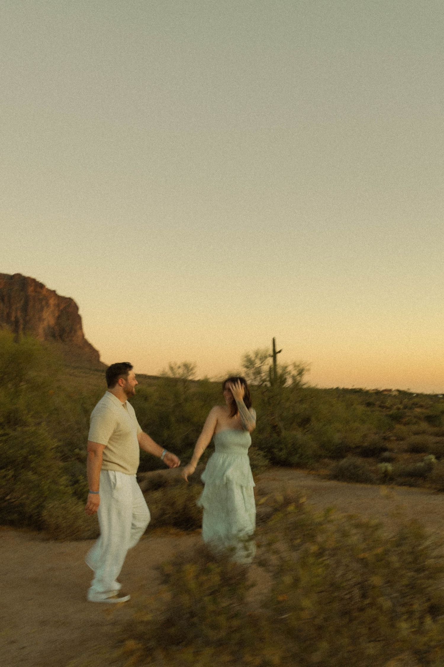 Couple holding hands strolling through Sonoran Desert at dusk with saguaro cactus and warm sunset sky backdrop.