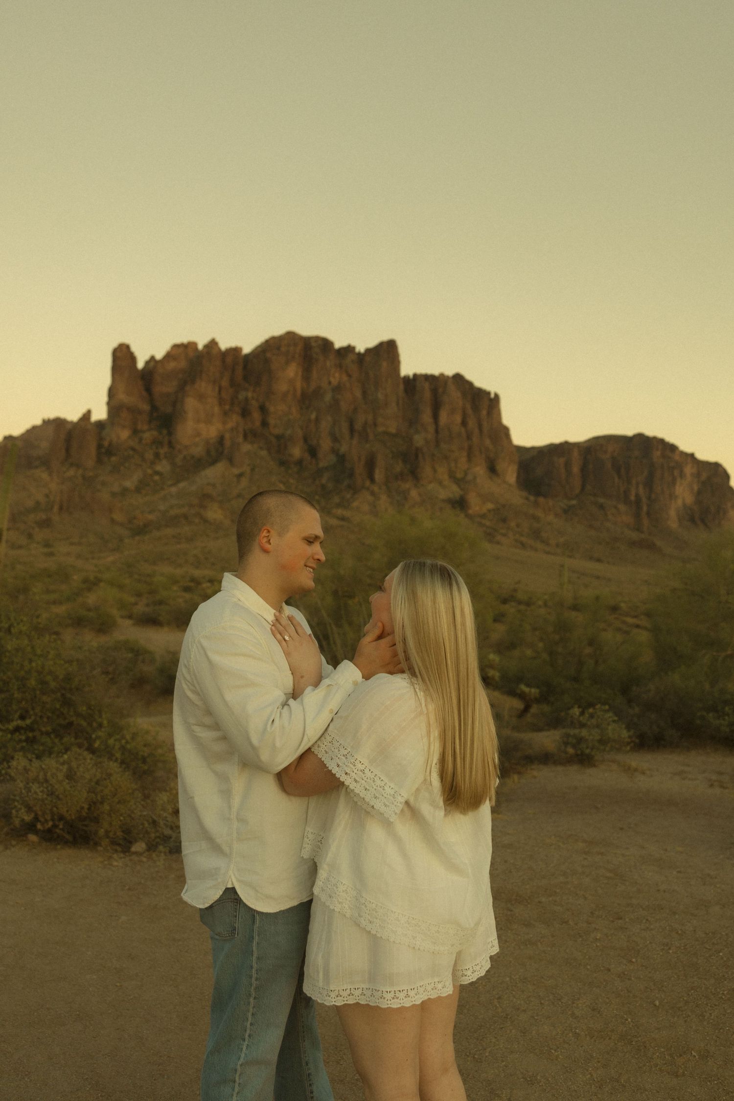 Couple in white embracing at sunset with Superstition Mountains in Arizona desert background.