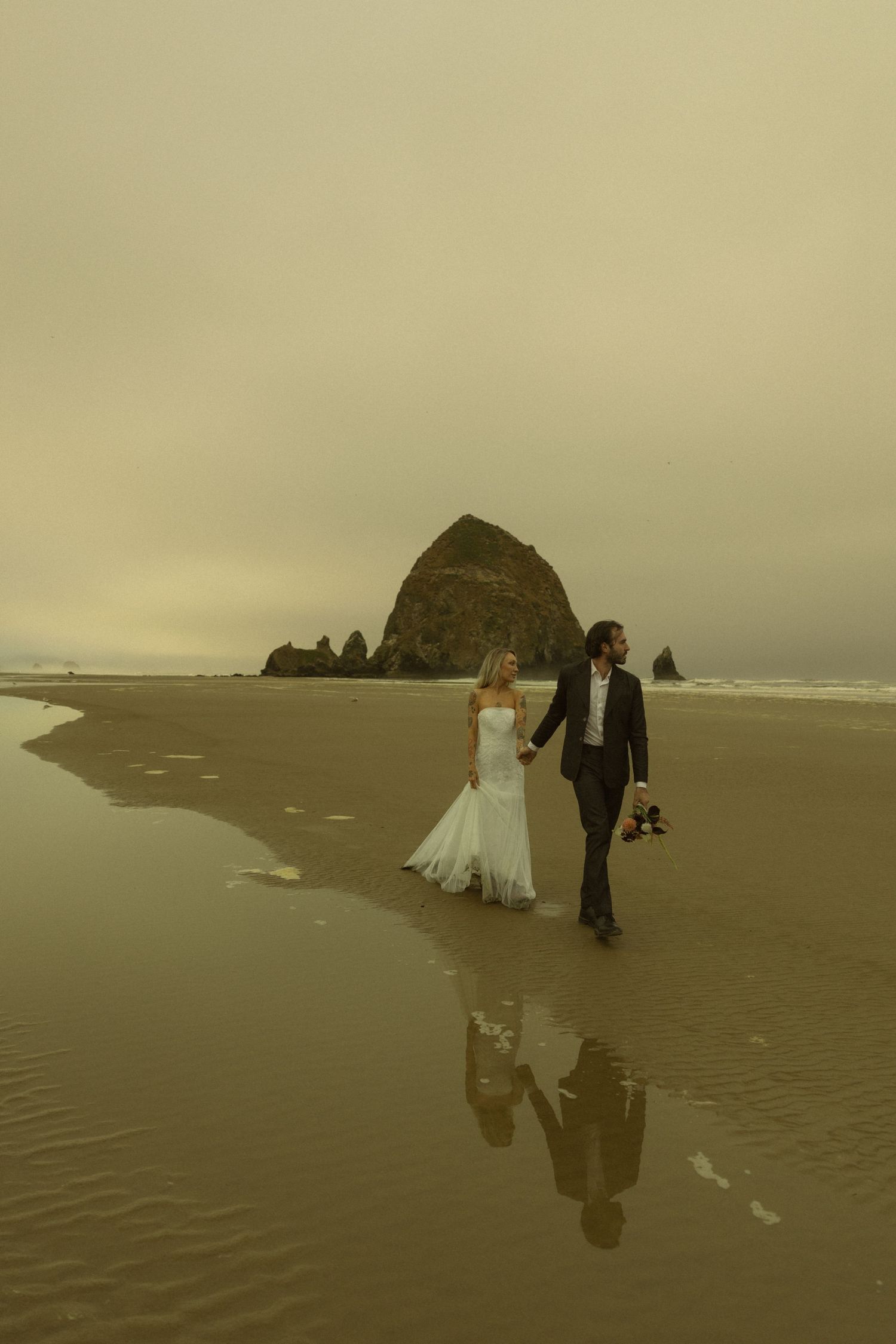 Newlywed couple walking hand in hand on a misty beach with Haystack Rock in the background at Cannon Beach, Oregon.