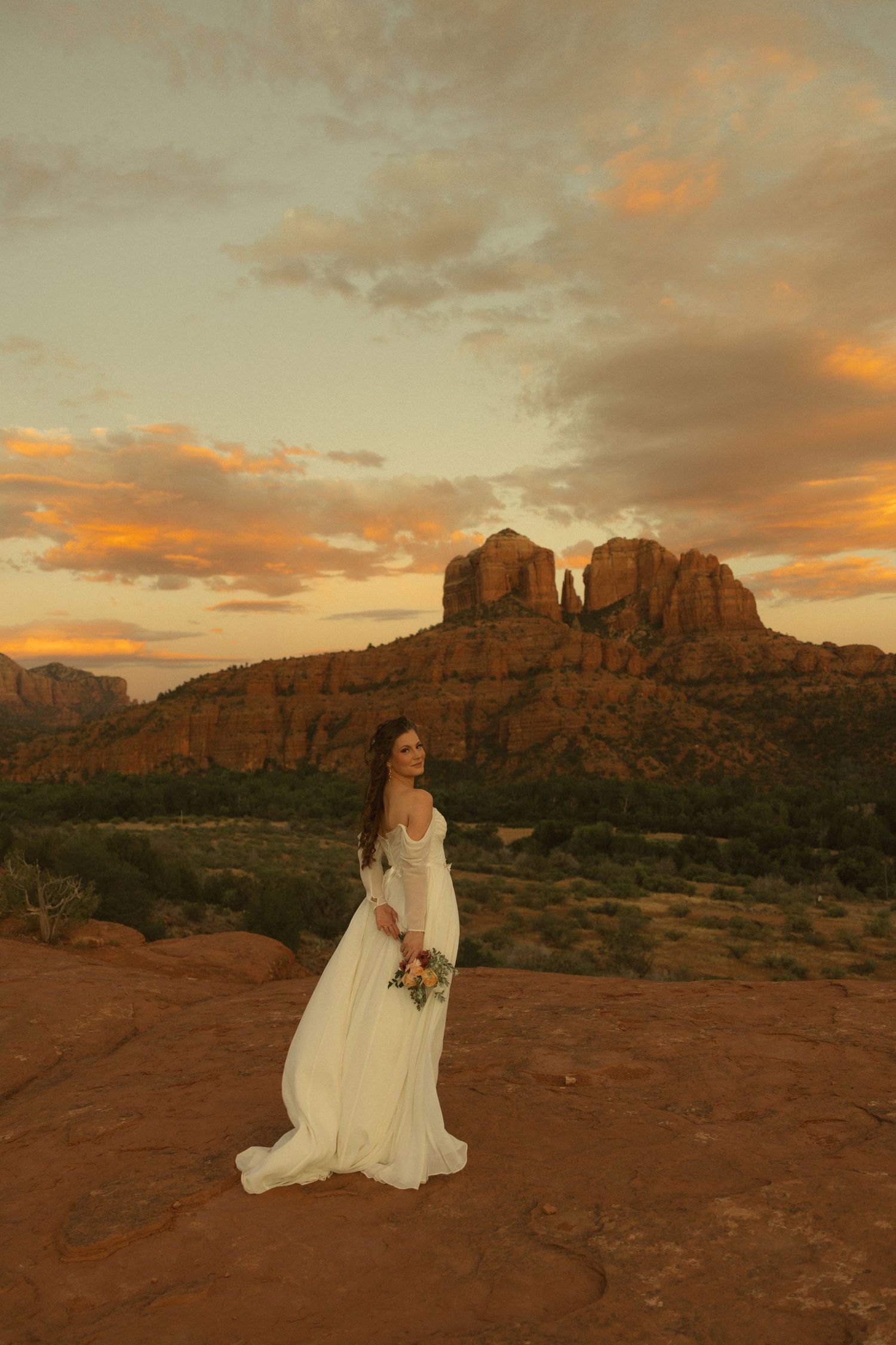 Bride in white lace gown holds bouquet while posing on red rocks at Cathedral Rock in Sedona at golden sunset.