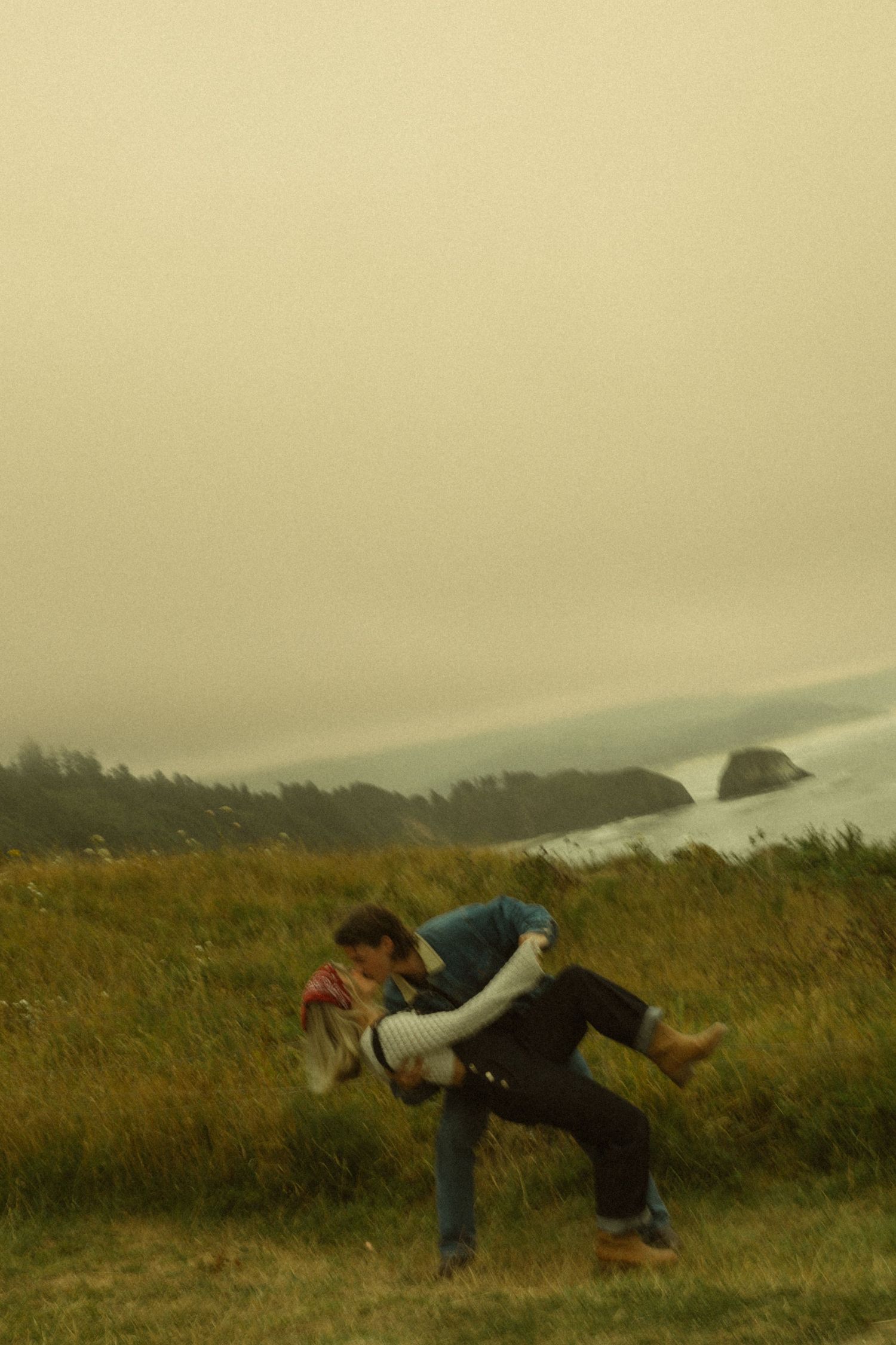 Couple playfully dancing on a grassy coastal bluff with misty ocean cliffs in the background.
