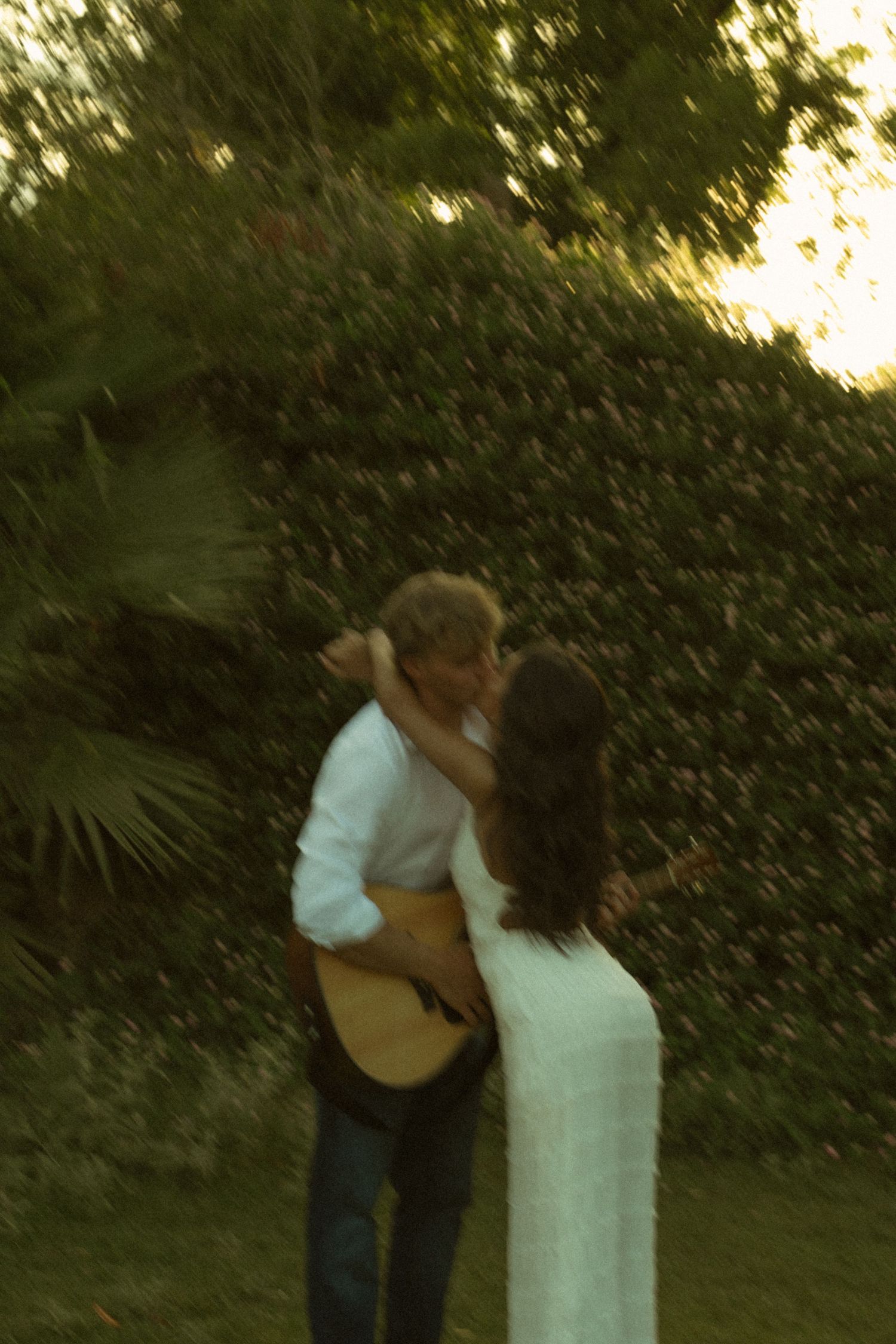 Couple sharing a tender kiss outdoors at dusk, man holding acoustic guitar in lush green garden setting.
