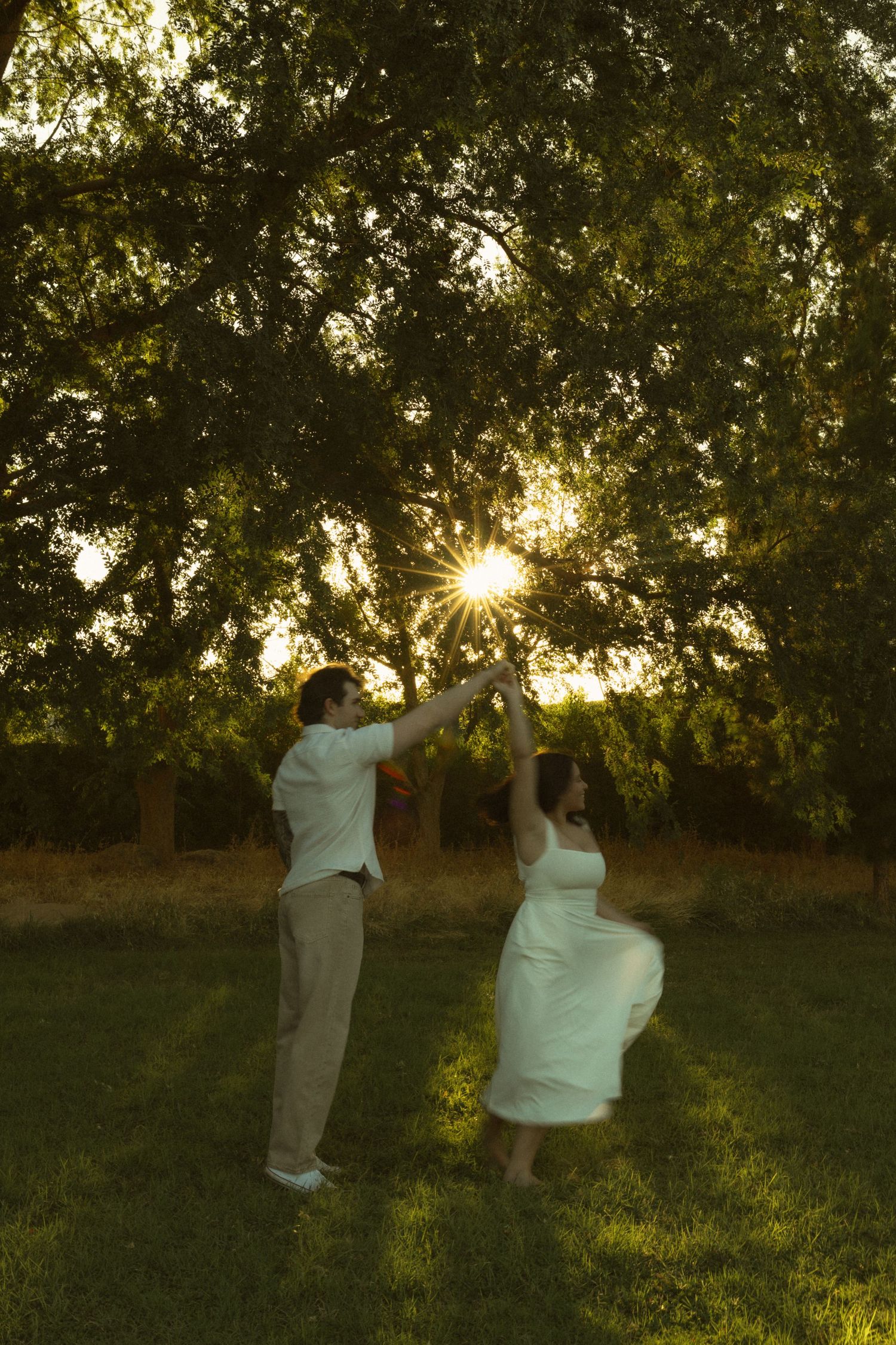 Couple dancing together at sunset under a large green tree on a grass lawn, golden light shining through the branches.