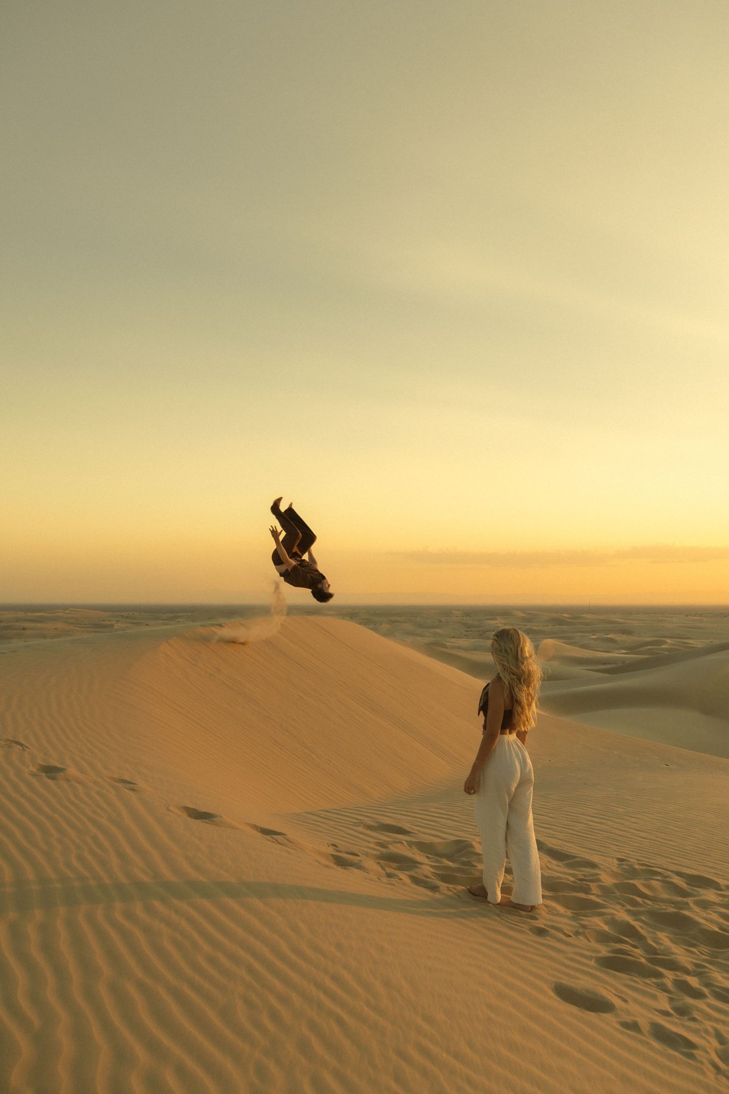 Woman in white jumpsuit watches a man doing a backflip off a sand dune at golden sunset.