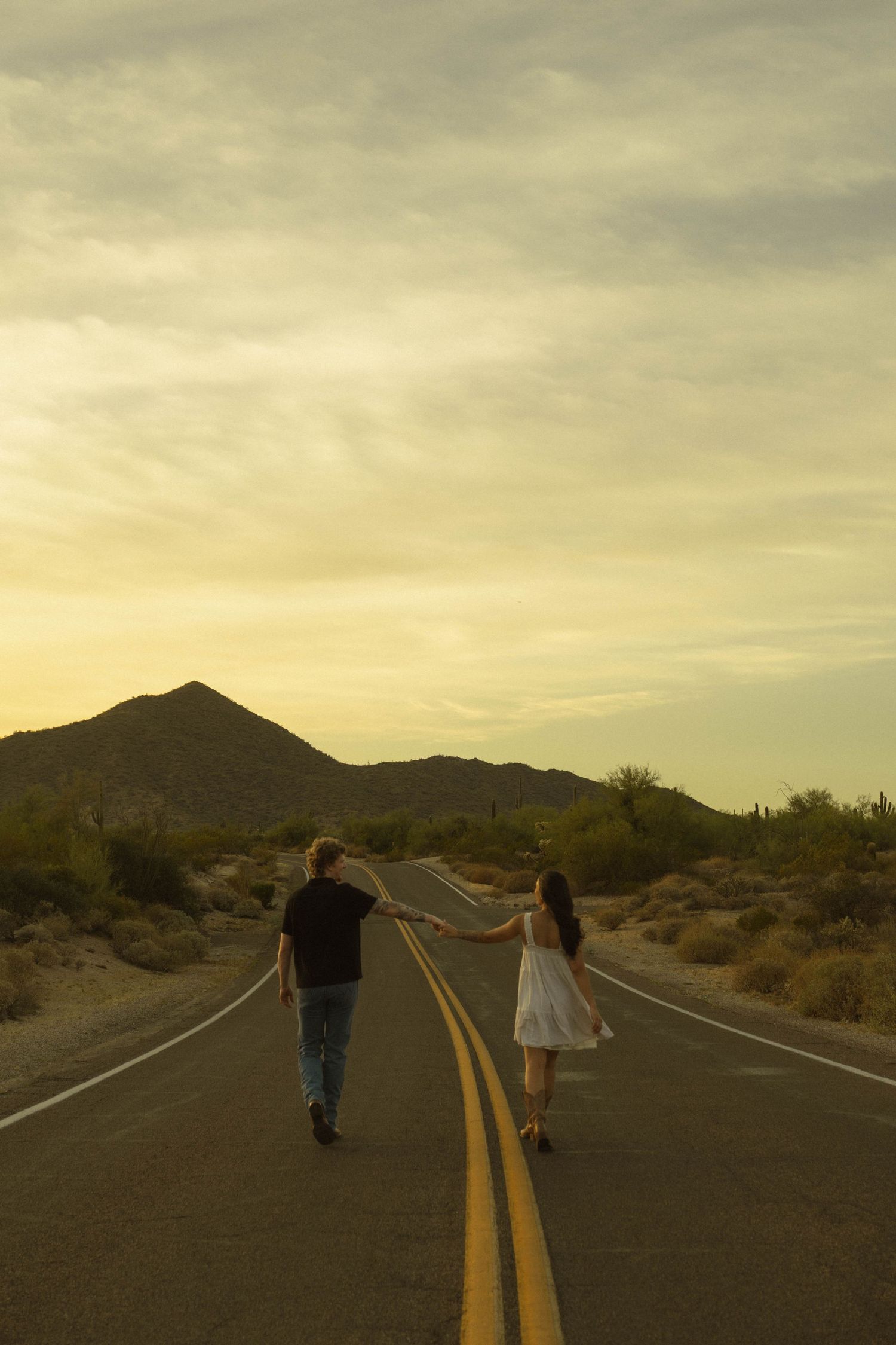 Couple holding hands walking down a desert road at sunset with mountains and cacti in the background.
