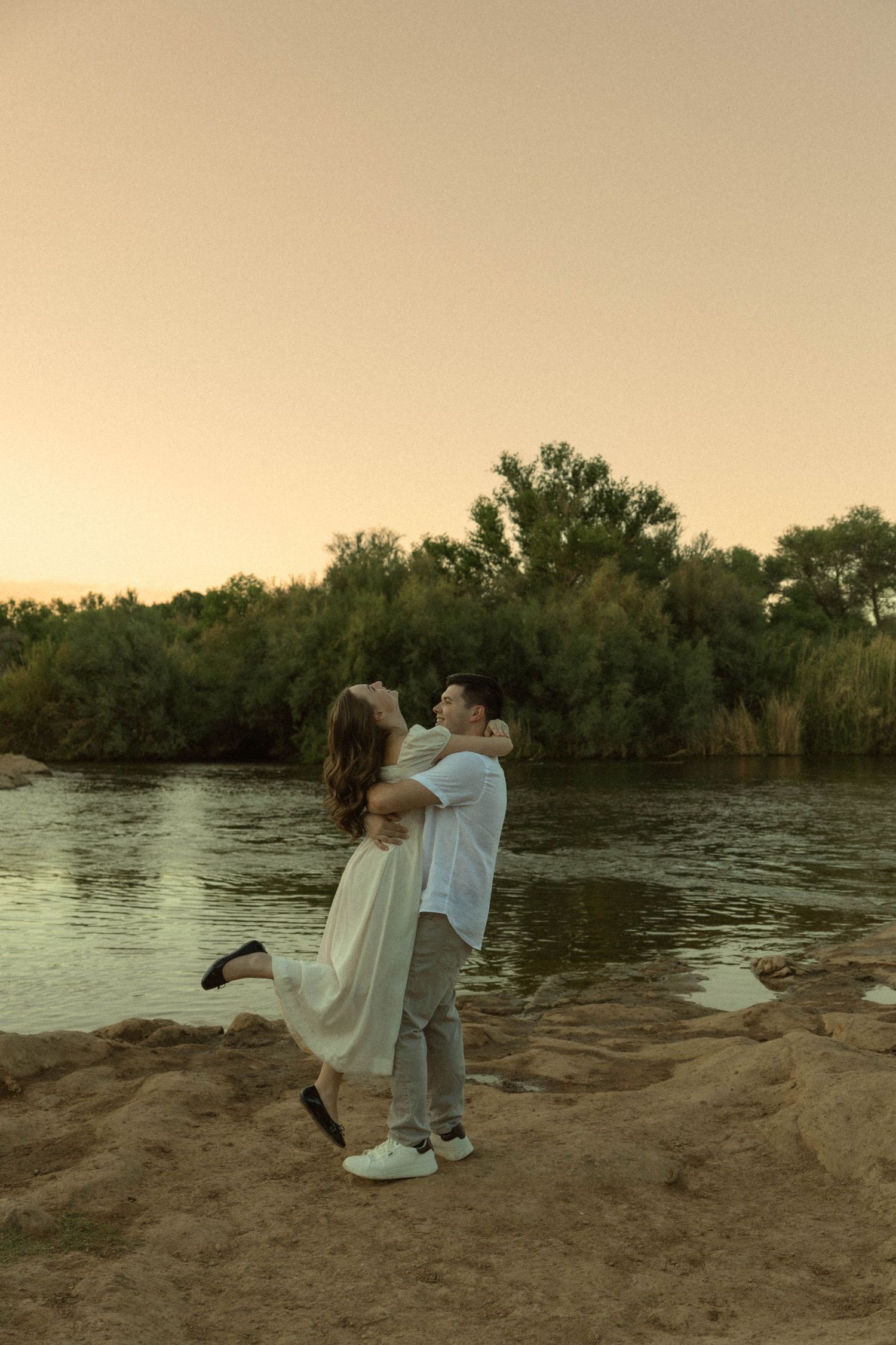 Couple embracing at sunset by a river, man lifting woman in white dress on sandy shore with lush green trees.