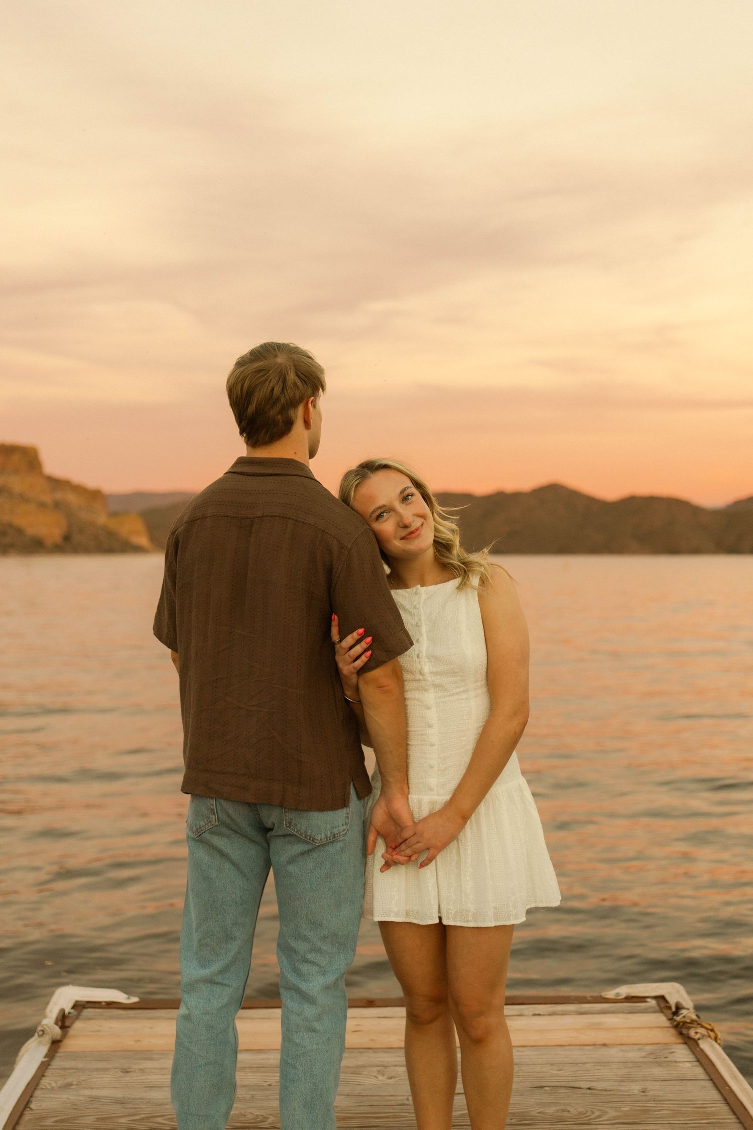 A couple shares a romantic moment on a wooden dock at sunset with mountains and water in the background.