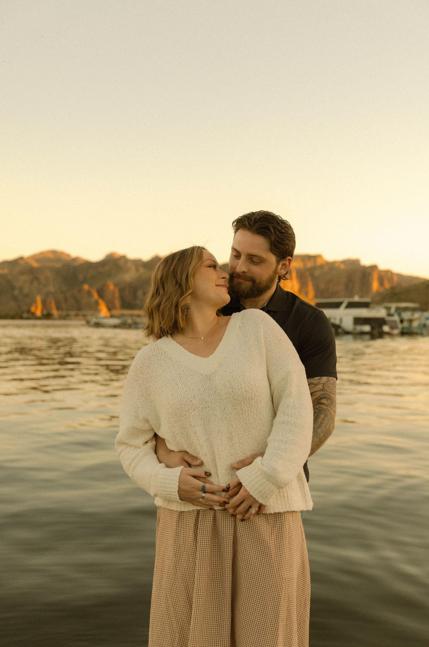 Romantic couple embraces at sunset by a lake with mountains in the background.