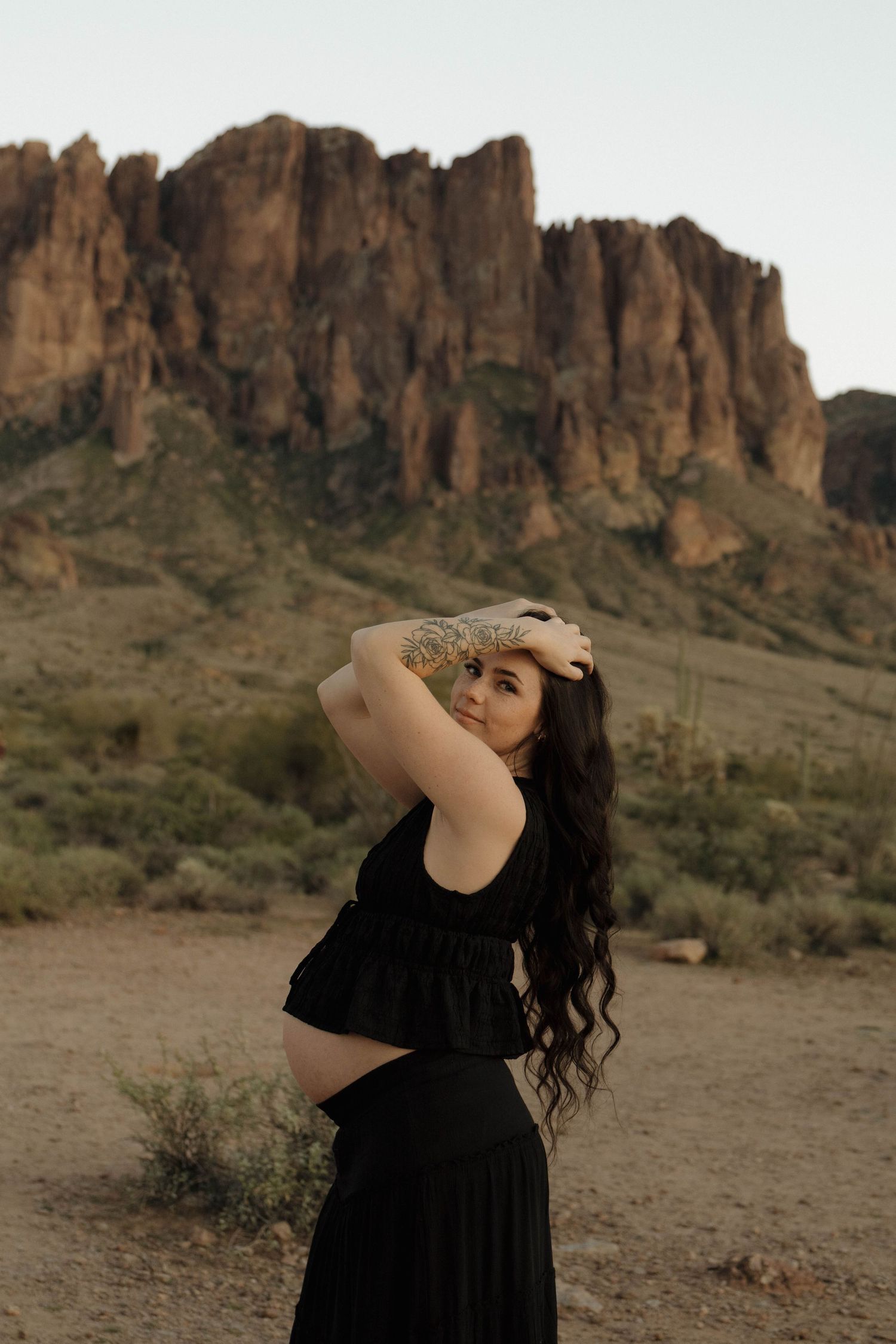 Woman in black dress poses gracefully against rugged desert mountain backdrop.