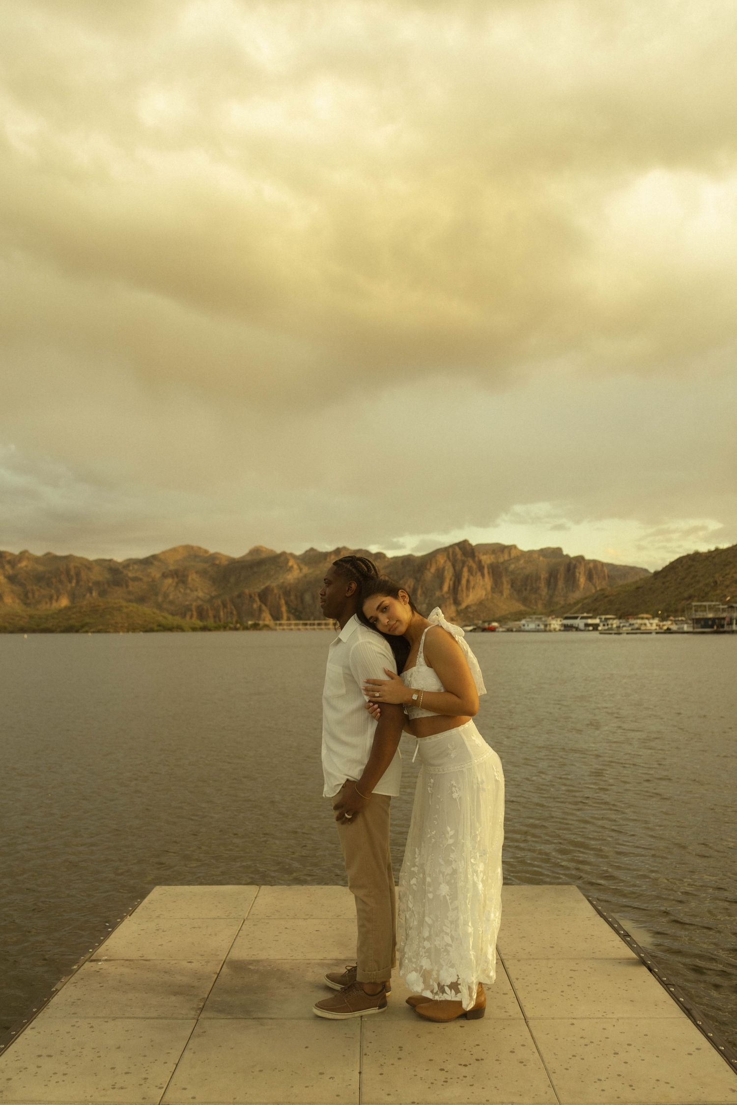 Couple sharing a tender embrace on a waterfront pier with scenic mountains under cloudy skies.