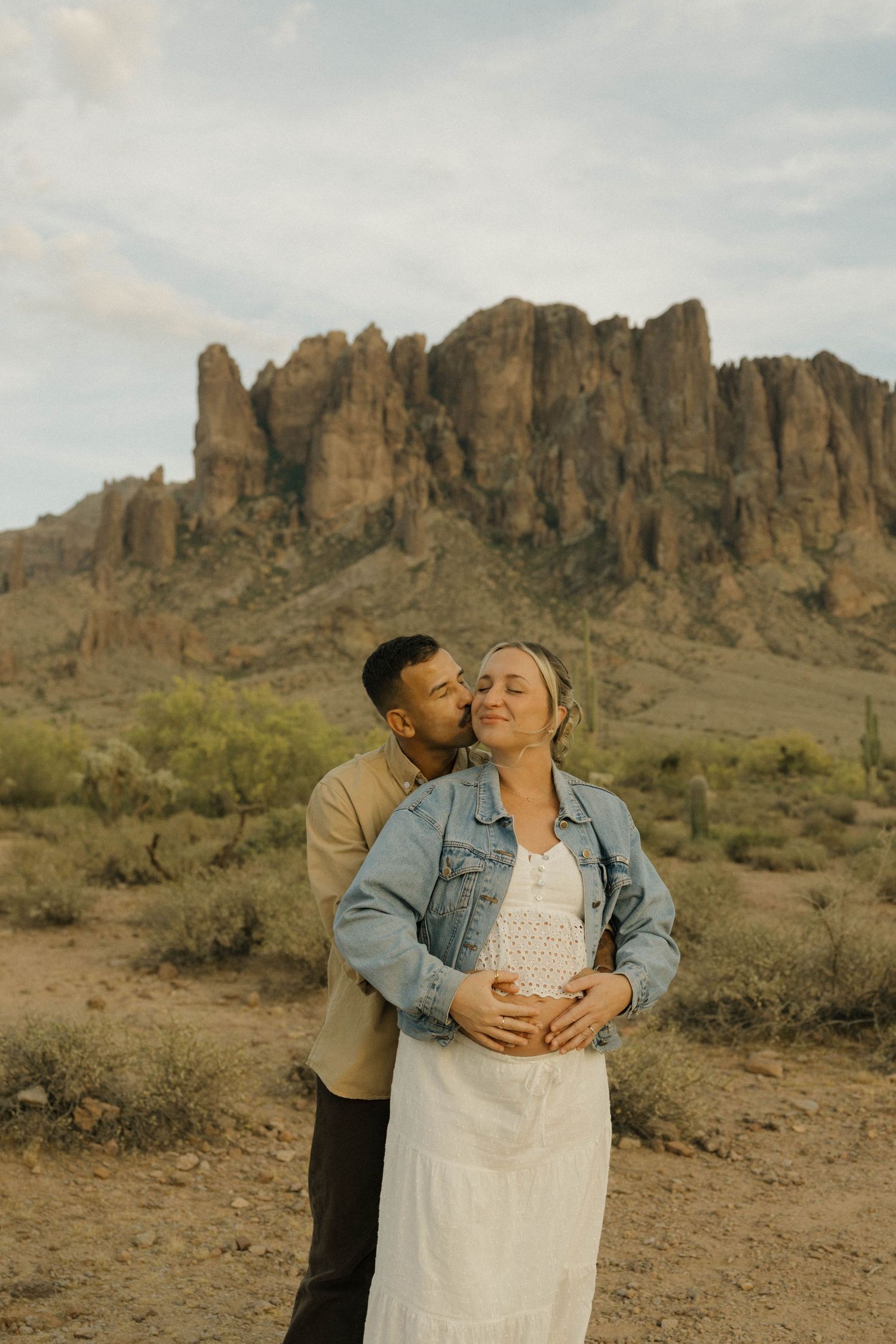 A couple embraces in the desert with Superstition Mountains backdrop, woman in white dress and denim jacket.