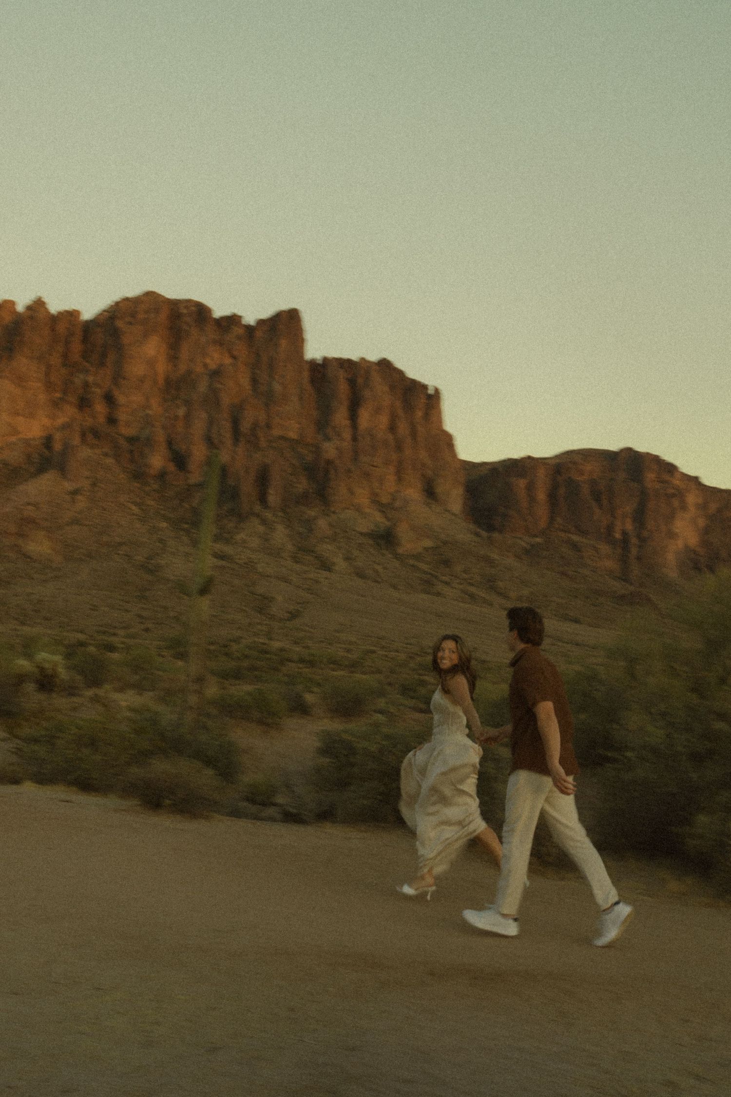 Couple holding hands walking in desert landscape with red rock mountains at golden hour sunset.