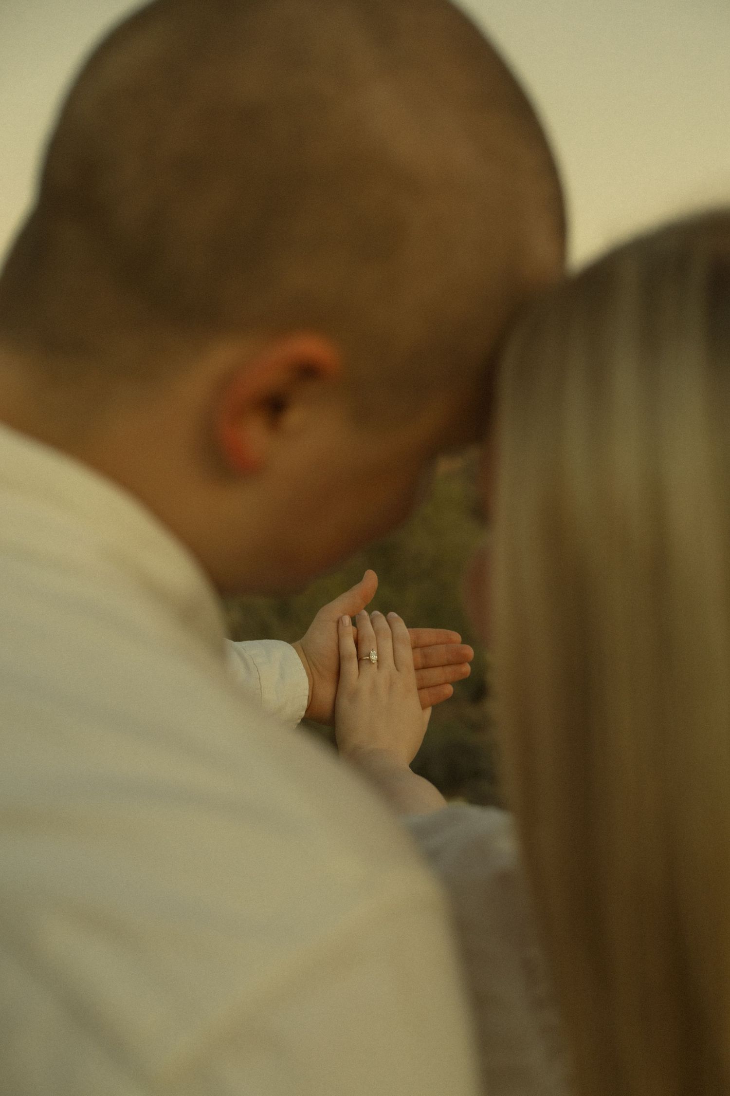Couple sharing a tender moment, woman displaying engagement ring on her hand while man kisses her forehead in warm light.