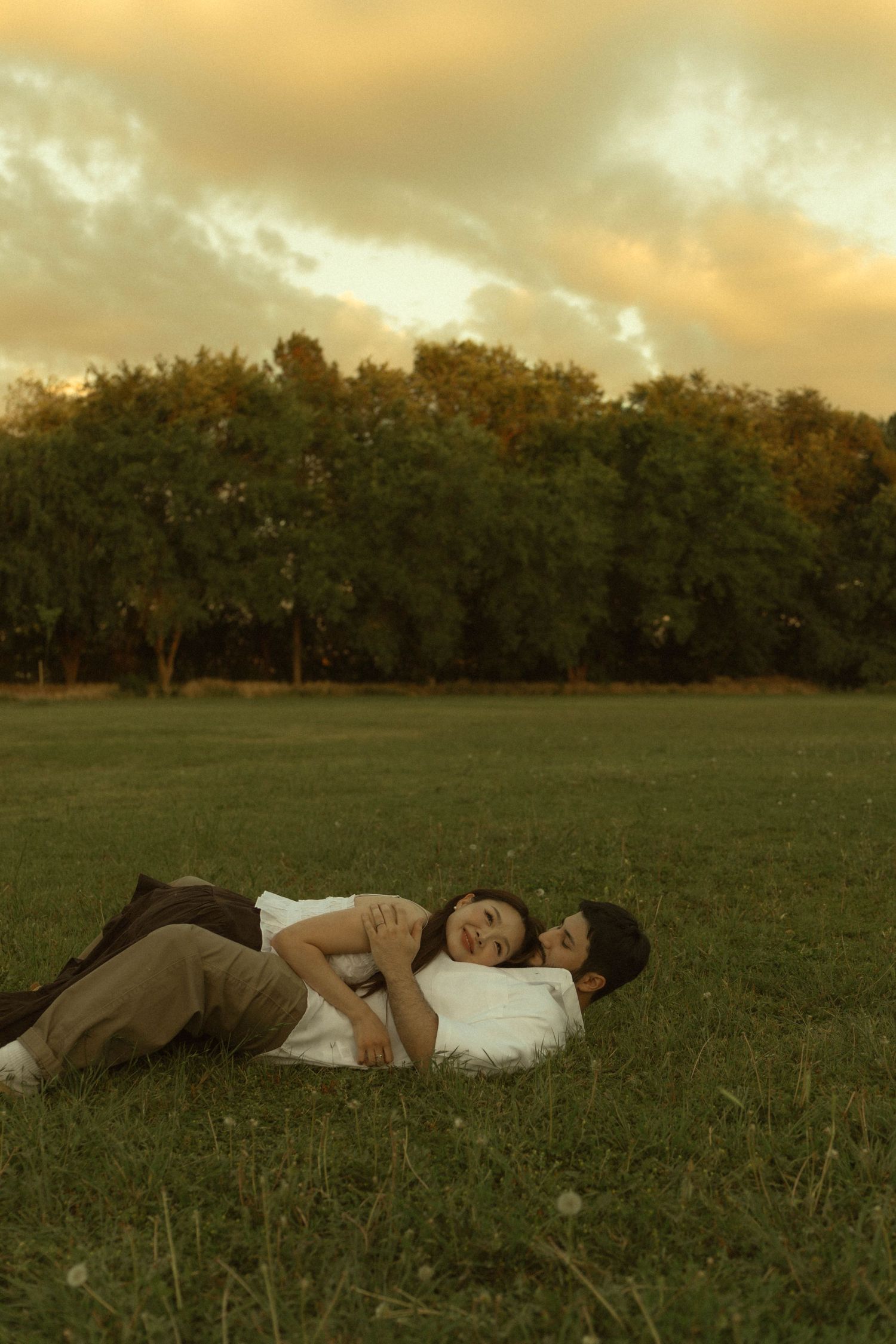 A happy couple lies together on green grass in a park under a dramatic golden cloudy sky.