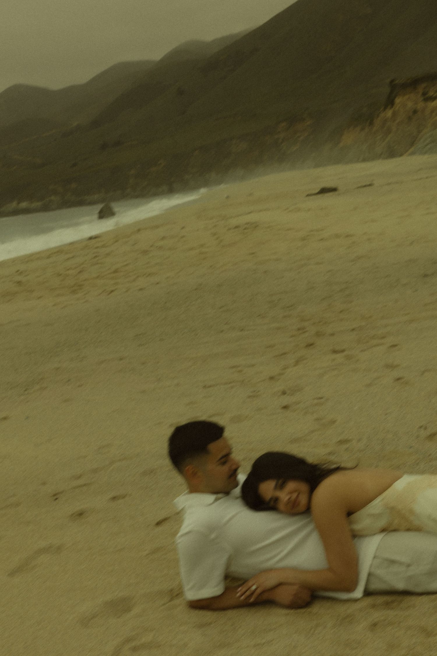 Couple lying together on sandy beach with coastal cliffs and ocean in the background.
