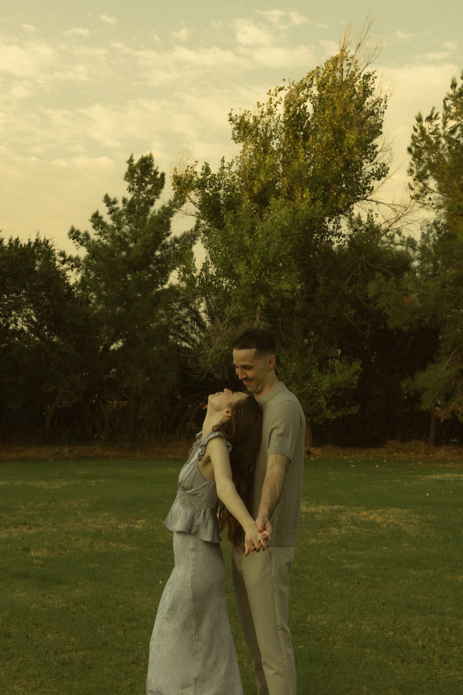 A couple holds hands and embraces on a green lawn surrounded by lush trees at golden hour.