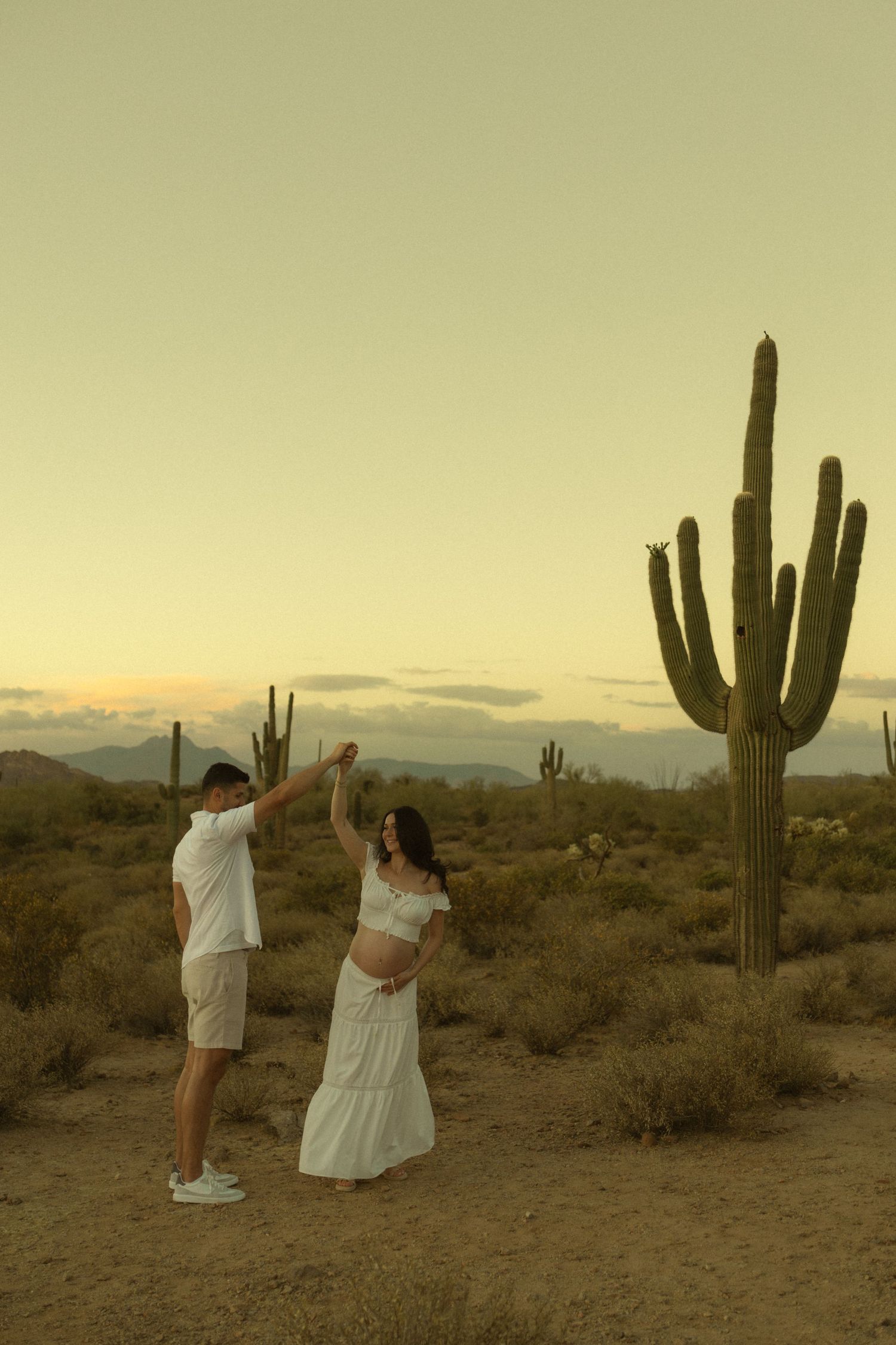 Couple dancing in white outfits at desert sunset with saguaro cactus, maternity photo shoot in Arizona.