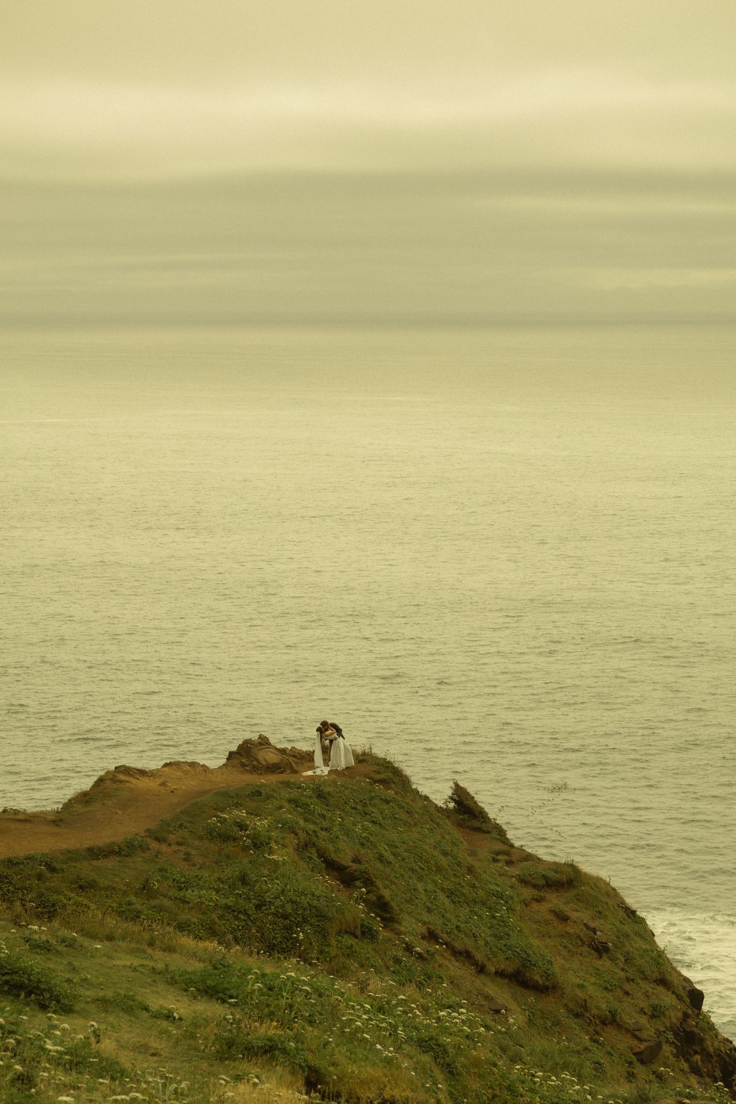Person sitting on a grassy coastal cliff overlooking a calm misty ocean under an overcast sky.