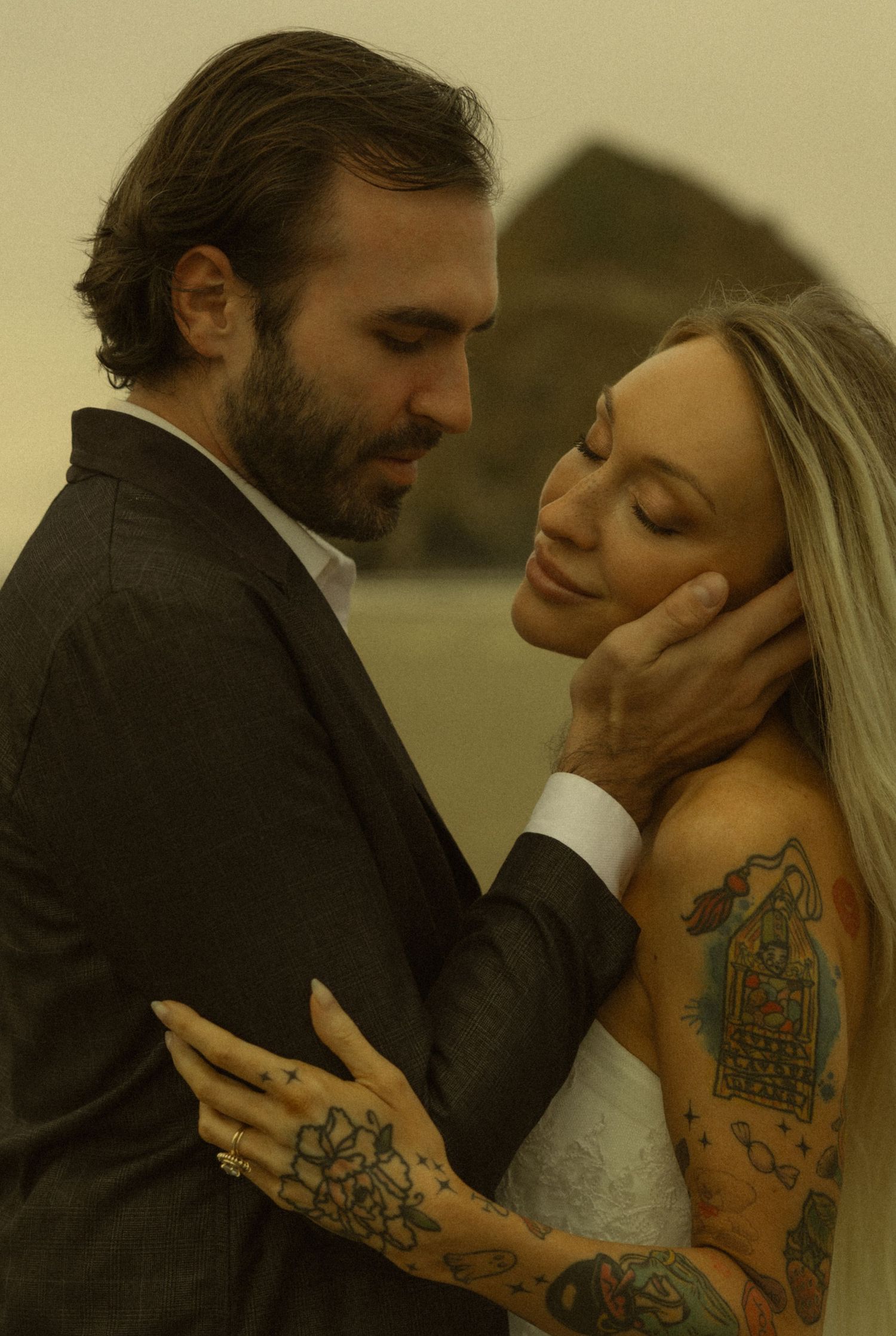 Couple in wedding attire sharing a romantic beach moment, woman with tattoos, man in dark suit.